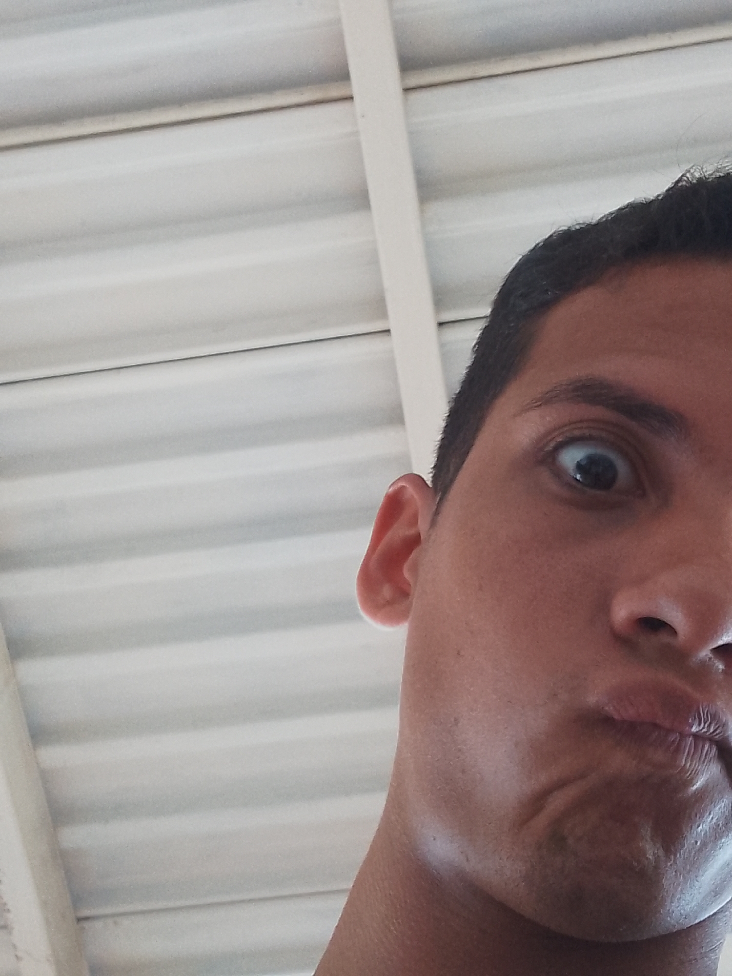 A close-up, low-angle photograph taken in El Espinal, Venezuela, captures the right side of an individual's face against a corrugated white ceiling. The person, appearing to be an adult male, has dark hair, a wide-open right eye with a visible pupil, and pursed lips. The facial expression conveys surprise or a humorous grimace, with the right ear also visible. The background consists of a uniformly white, horizontally ribbed ceiling structure, likely made of metal or PVC panels, with a faint vertical white support beam present on the far left. The lighting is diffused, illuminating the subject and background evenly.