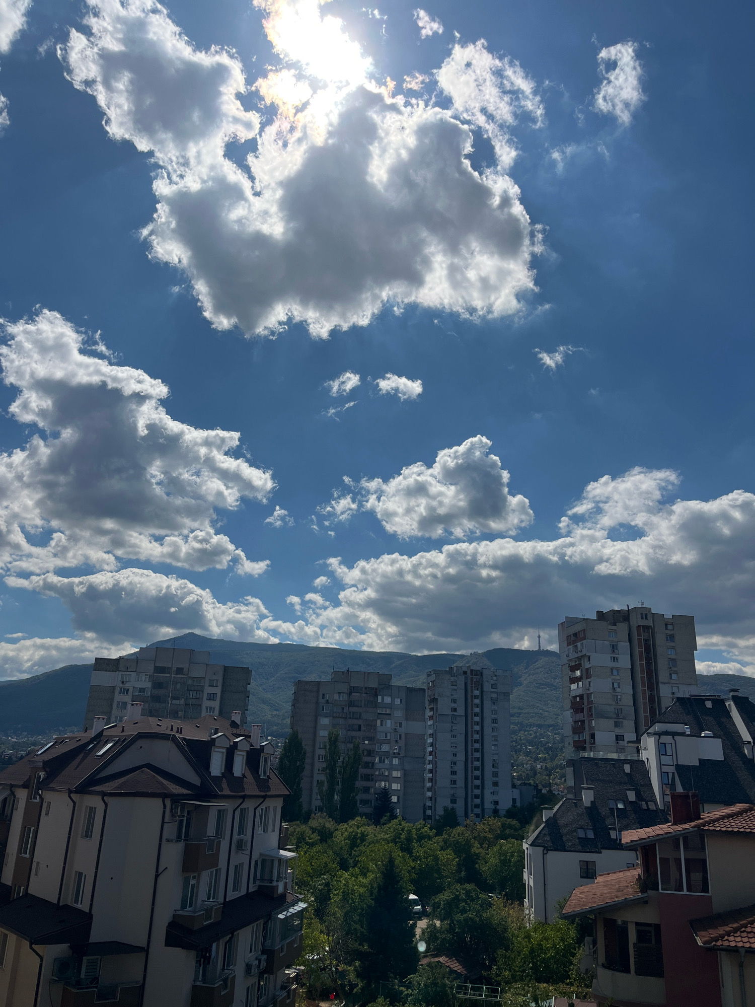 The image depicts a cityscape from an elevated perspective in Sofia, Bulgaria, likely captured during daytime given the bright sunlight. The sky is a vibrant blue, dotted with numerous fluffy white clouds. The sun is partially obscured by a large cloud formation in the upper center, casting rays of light downwards.
In the foreground and mid-ground, several multi-story buildings are visible. One building in the immediate foreground, on the left, is lighter in color with a brown roof and multiple windows and dormers. To its right, a cluster of taller, grey apartment buildings dominates the middle ground. Further back, another tall, reddish-brown building stands out on the right. Behind these buildings, a dense canopy of green trees is present, suggesting a park or residential area with abundant vegetation.
In the background, rolling hills covered in greenery rise, with the faint outline of mountains beyond. A tall tower, possibly a telecommunications tower, is visible on one of the distant hills. The overall impression is a view of an urban environment nestled against a natural landscape under a partly cloudy sky. No people or specific actions are discernible in the image.