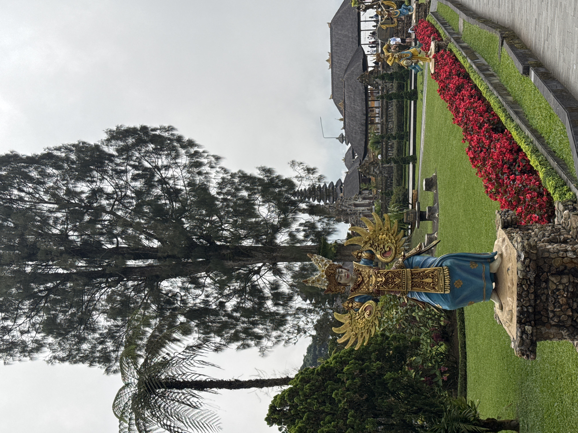 The image depicts a scene from what appears to be a temple or garden complex, possibly in Bali, Indonesia, but the specific location is unknown. A statue of a deity or mythical figure is in the foreground, painted in blue and gold and adorned with ornate details. The statue is set upon a stone base. Behind the statue, there is a large tree with dense foliage. A pathway lined with red flowers and manicured greenery runs through the scene, leading towards a multi-tiered structure with traditional Balinese architecture, including a thatched roof. People are visible in the background, suggesting the complex is open to the public. The sky is overcast.
