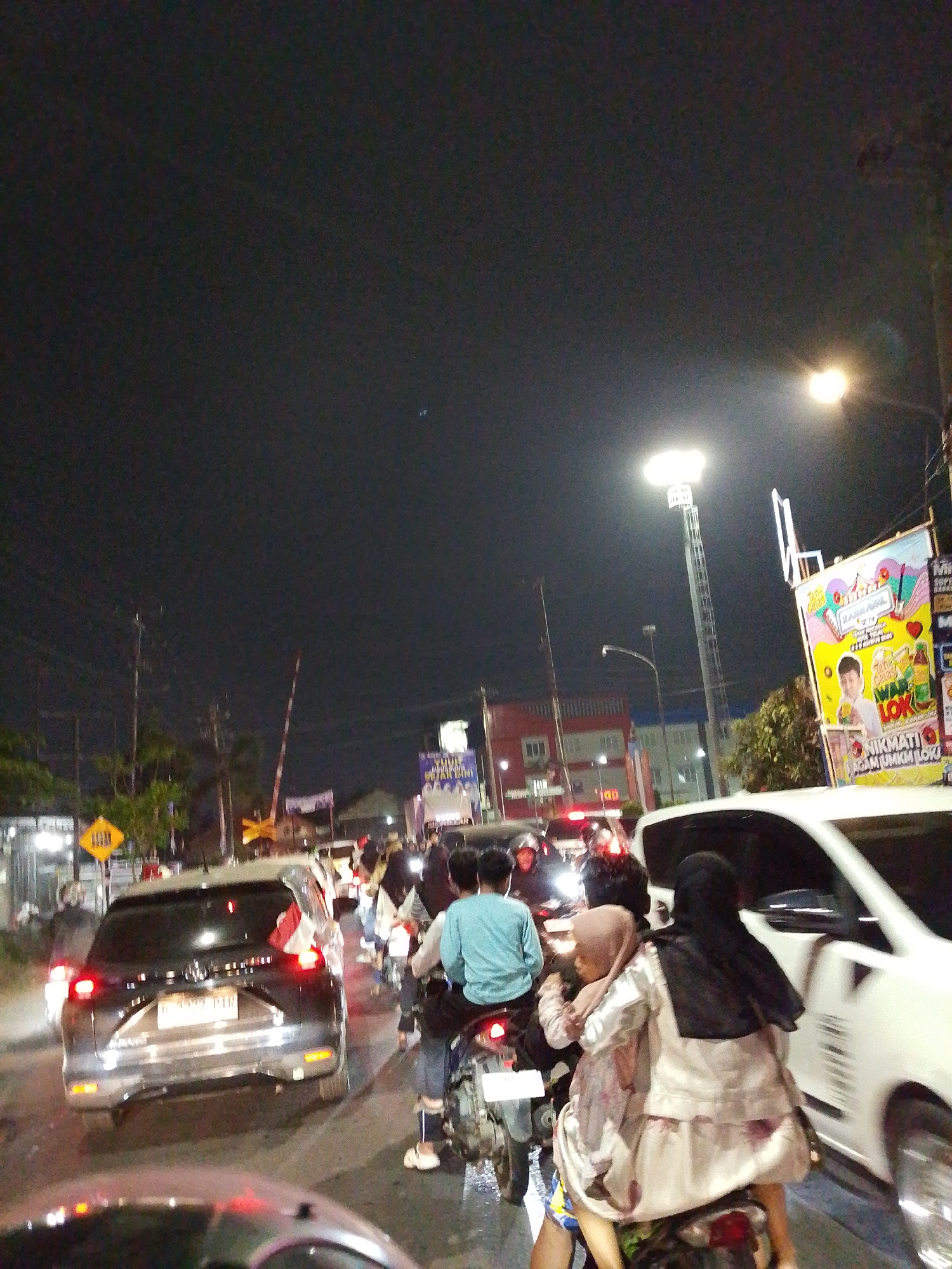 The image depicts a traffic jam at night in Tegal, Indonesia. A traffic light appears to be malfunctioning, with a red and white barrier lowered across the road, indicating a train crossing. Numerous motorcycles and cars are stopped on the road. On the right side of the image, a large billboard advertises a local business. The overall atmosphere is one of congestion and potential delay due to the train crossing.