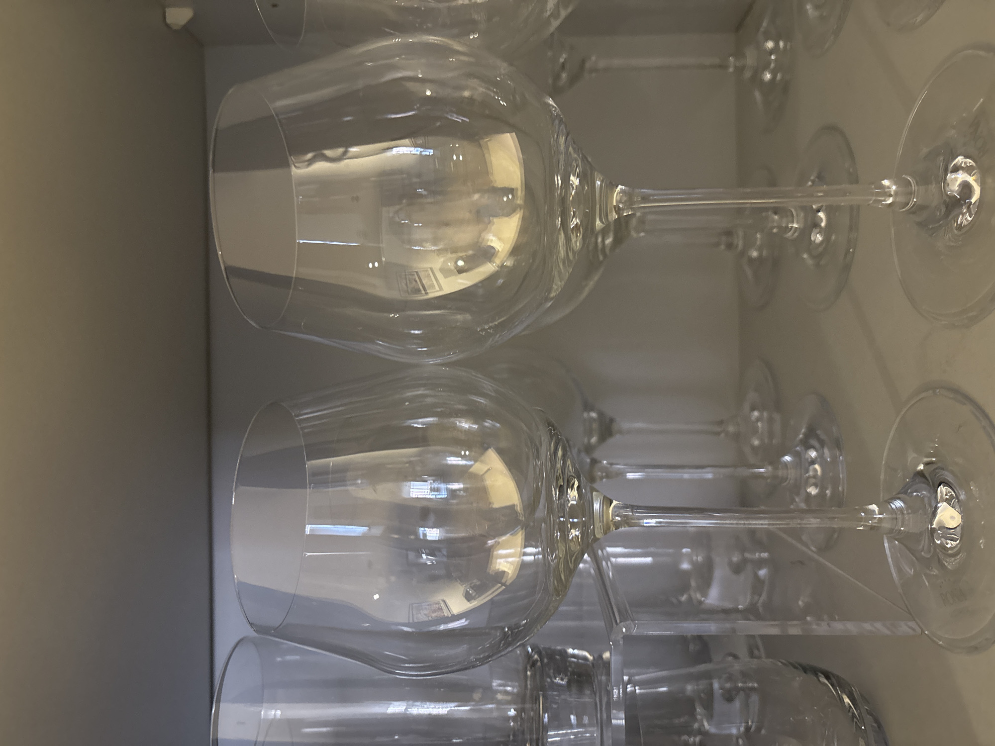 The image shows a close-up, eye-level view of several empty, clear glass wine glasses arranged on a light-colored shelf. The glasses are the primary focus, with two prominent ones in the foreground. The glass on the left has a round bowl and a slender stem, while the glass on the right has a wider, more bulbous bowl. Reflections of what appear to be room interior elements, including lighting and possibly framed pictures, are visible on the surfaces of the glasses. In the background, other glasses and glassware are partially visible, suggesting a collection or storage area. The overall environment appears to be indoors, possibly a kitchen or display cabinet, with a neutral, grey background above the shelf. There are no people or animals depicted in the image. The scene is static and lacks any discernible action. The lighting is ambient, creating some glare and reflections on the glassware. The image was taken in Dubai, United Arab Emirates.