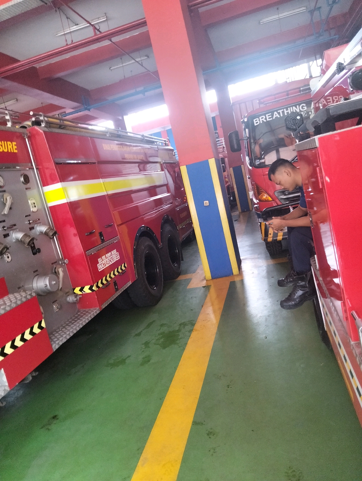 The image depicts the interior of a fire station in South Jakarta, Indonesia. Two red fire trucks are parked side-by-side. The fire truck on the left is closer to the camera, showing its side with equipment and hoses. The fire truck on the right is partially visible, with its windshield displaying the text "BREATHING". A male firefighter, wearing a dark blue uniform and black boots, is seated on the ground in front of the fire truck on the right, looking down at his phone. The fire station has a green floor with yellow markings. Red beams and a white ceiling are visible above. The overall scene suggests a moment of downtime or waiting at the fire station.