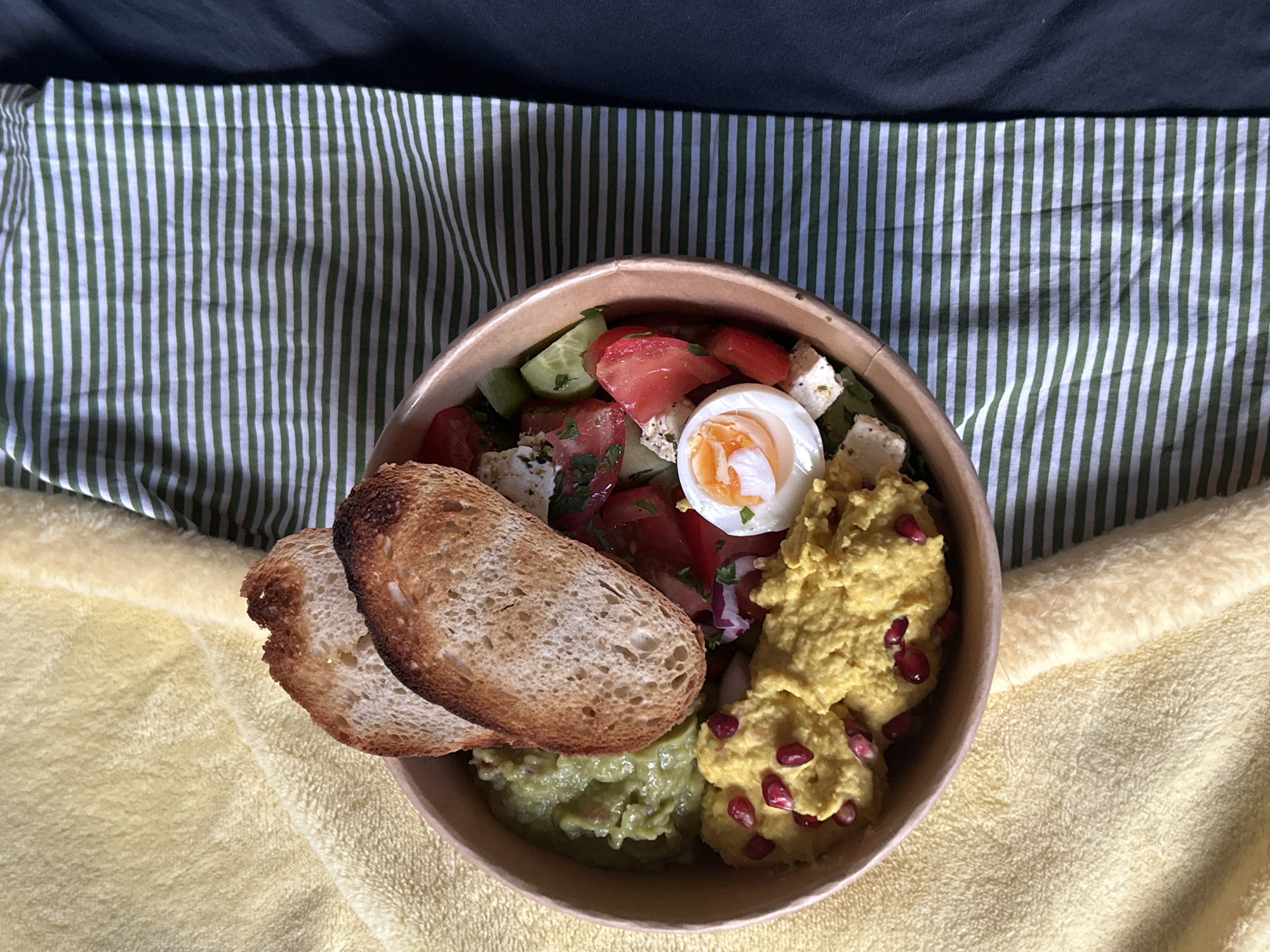 The scene depicts a single light brown, round paper bowl containing a varied meal, positioned on a surface within the city of Sofia, Bulgaria. The bowl's contents include a portion of light green mashed avocado in the top left quadrant. Two slices of toasted bread, exhibiting browned edges, are placed vertically above the avocado. To the right, a salad mixture is present, comprising diced red tomatoes, sliced green cucumbers, and crumbled white feta cheese, garnished with green herbs. A halved hard-boiled egg, revealing a firm orange yolk, is situated below the salad. In the bottom left, a yellow, textured paste, topped with several red pomegranate seeds, completes the meal. The bowl rests on a textured, soft yellow fabric that covers the left side of the visible surface. To the right of the yellow fabric, a white textile with evenly spaced horizontal green stripes is visible. The far right background displays a solid dark grey fabric. No individuals, actions, or interactions are observable in the image.