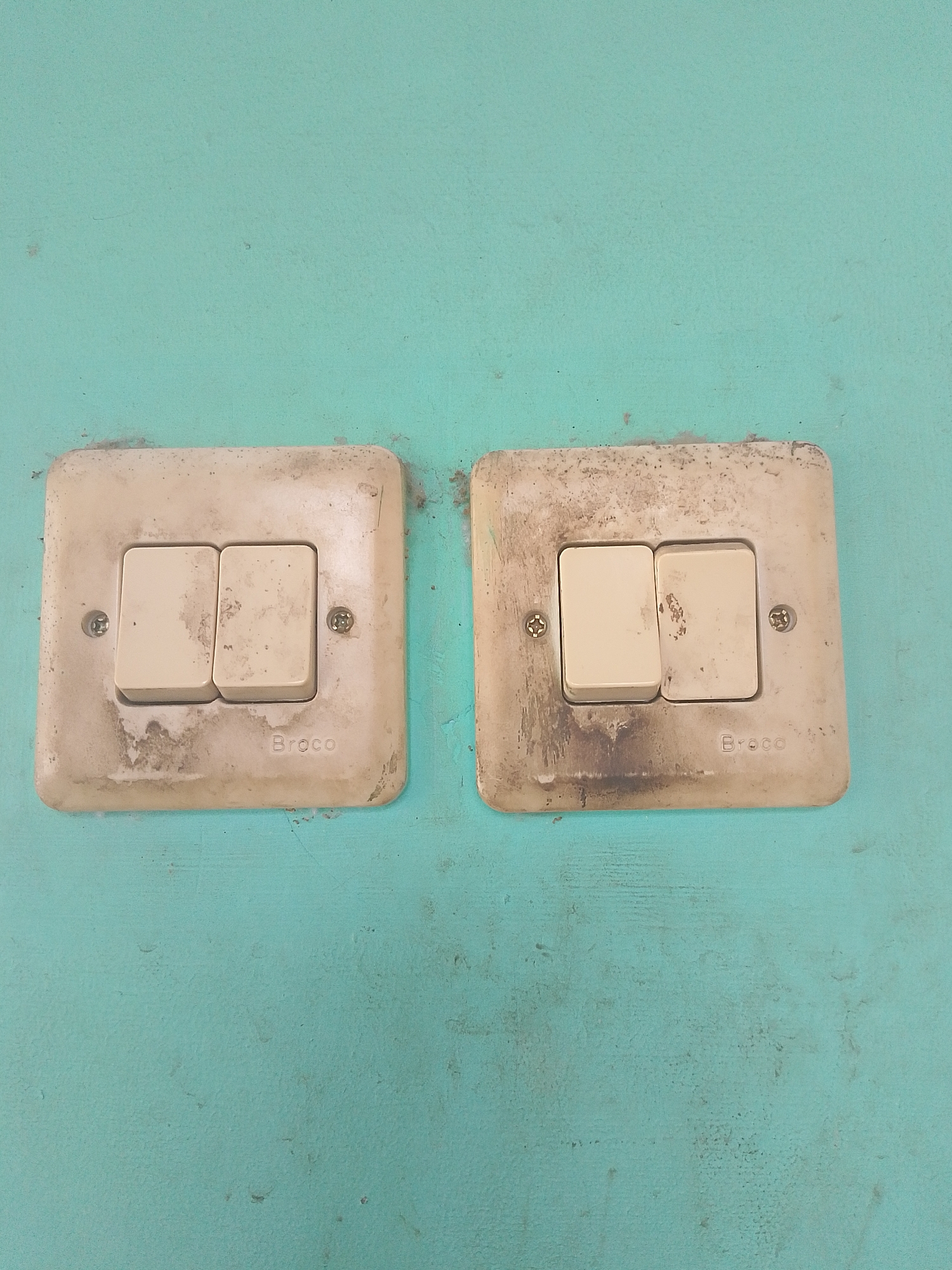 The image shows two light switches mounted on a turquoise wall. Each switch has two beige buttons. The switch on the left has "Broco" written on the bottom right of its plate. The switch on the right also has "Broco" written on the bottom right of its plate. Both switch plates are discolored and appear dirty, with some dark smudges and dirt. The wall itself has a textured surface with some minor imperfections and dark specks. There are no people or actions depicted in the image. The overall impression is of an outdoor or utilitarian indoor setting, possibly in a public area given the brand name visible on the switches, which are common in Indonesia. The location in Situwangi, Indonesia, suggests this is a snapshot of everyday electrical fixtures in that region.