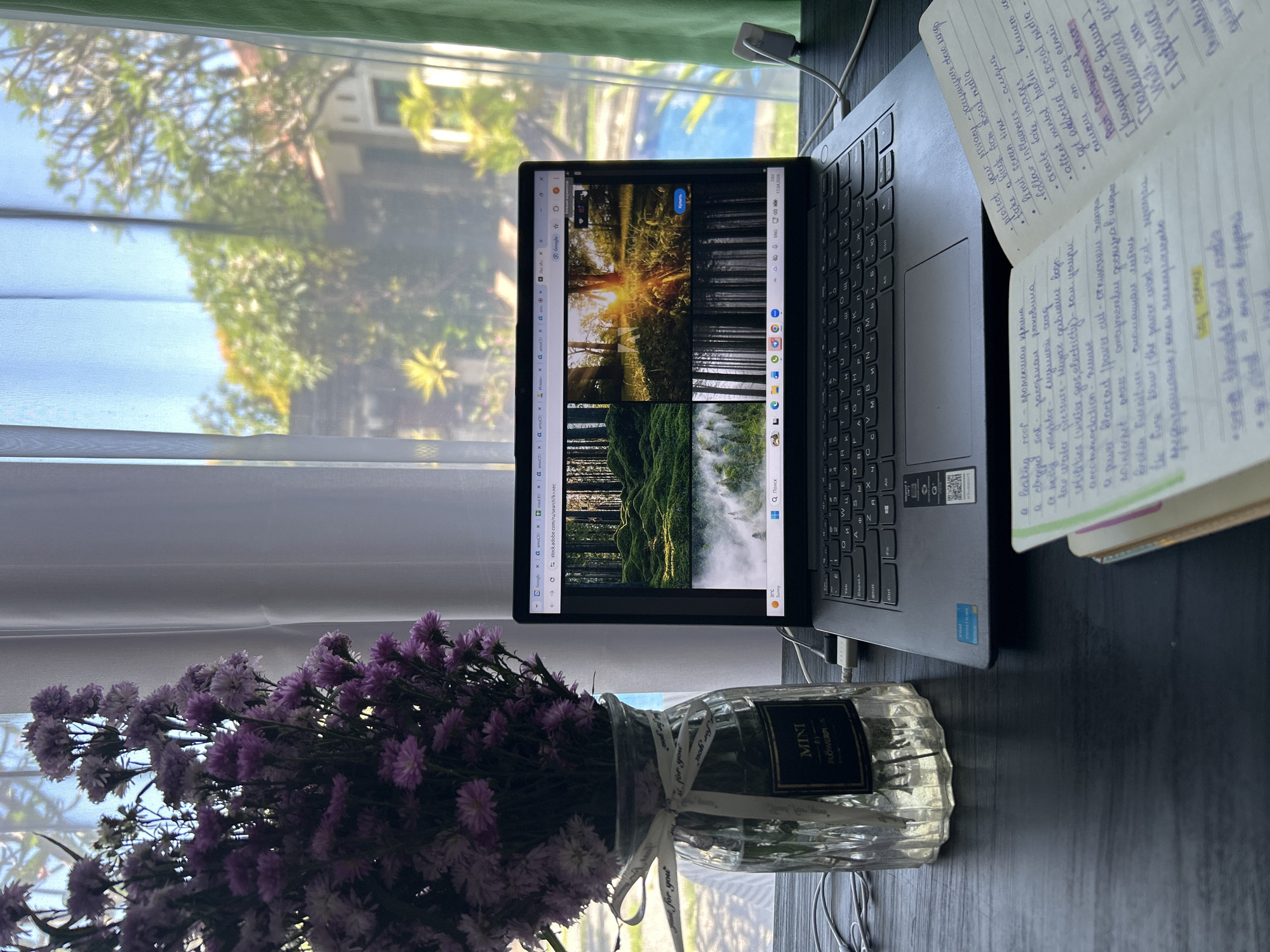 The image displays an indoor scene by a window, featuring a laptop, an open notebook, and a vase of purple flowers. Bright natural light illuminates the setting, suggesting daytime.
On the left, a clear glass vase holds a bouquet of small purple flowers. The vase has a label reading "MINI FLOWERPLUS" and a white ribbon tied around it, imprinted with "for you".
A dark gray laptop, likely a Lenovo with "Intel Core i5" and "Windows" stickers visible, is open on a dark desk. Its screen shows a web browser with multiple tabs. The active tab displays Google search results for nature images, specifically from "stock.adobe.com/ru/search?k=лес" (forest in Russian). The images include a sunrise in a forest, dense dark woods, and a misty forest scene. Visible text on the screen includes "Google", "Поиск" (Search), "Крепить" (Attach/Pin), "ENG", and "31°C", indicating the current temperature. Other tabs show repeated instances of "amox" and "Исман".
To the right of the laptop, an open notebook contains handwritten notes, primarily in English with Russian translations. The left page lists phrases such as "protect your privacy", "limit screen time", "follow influencers", "affect mental health", and "get addicted to social media". The right page details household problems like "a leaking roof - протекшая крыша", "low water pressure - низкое давление воды", "a power blackout (power cut) - отключение электричества", and "no internet access - отсутствие доступа в интернет". The heading "Past Continuous tenses" is also present.
Through the window, a bright outdoor view is visible, partially obscured by sheer white curtains. It shows green foliage from trees, a blue sky, and a glimpse of a building. The sunny, warm outdoor environment is consistent with the location context of Kuta Selatan, Indonesia.