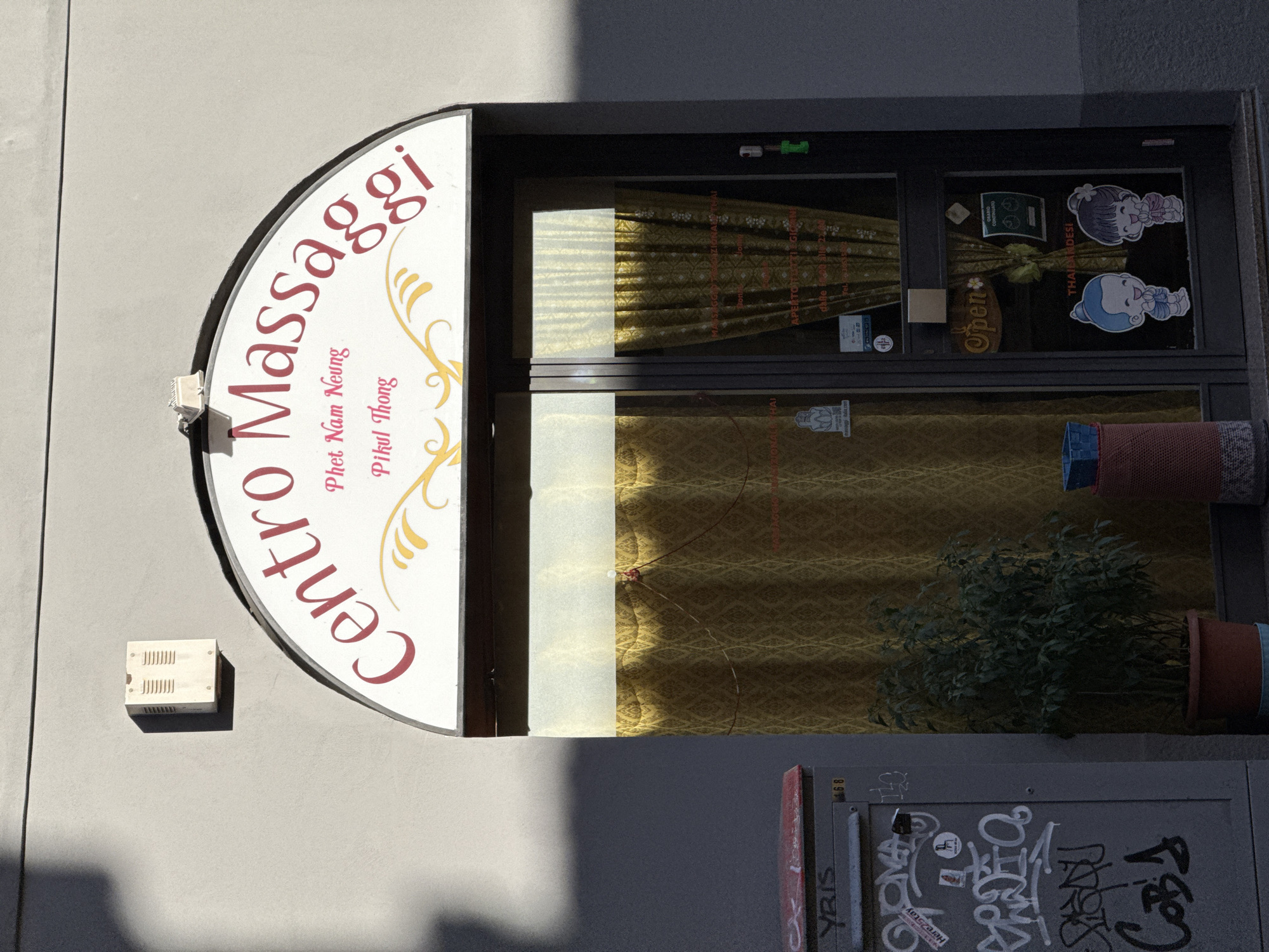 This is a medium-shot, outdoor, eye-level, full-color photograph of the entrance to a Thai massage business in Milan, Italy, with a focus on the signage and storefront. The building's facade is a neutral gray, with a shadow cast from the left. Above the entrance, an arched sign reads "Centro Massaggi" in red lettering with yellow decorative elements. Below this, smaller text states "Phet Nam Neung" and "Pikul Thong". The storefront consists of a double glass door with yellow patterned curtains visible behind it. On the left side of the door, there is a gray mailbox with graffiti and stickers. To the right of the door, a small green sticker is affixed to the door frame. Below the "Open" sign, there are two cartoon stickers of children in traditional Thai clothing. A potted plant sits to the left of the door. The overall scene suggests a commercial establishment in an urban setting.