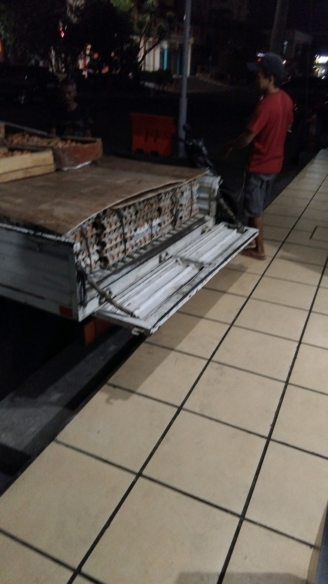 In Pekalongan, Indonesia, at night, a person wearing a red t-shirt and gray shorts stands on a sidewalk next to a truck bed loaded with egg cartons. The truck bed's tailgate is down, revealing white plastic interior. The sidewalk is paved with light-colored square tiles with dark grout lines. In the background, trees and buildings are visible, along with an orange traffic barrier. The scene suggests a commercial or logistical activity taking place in an urban environment.