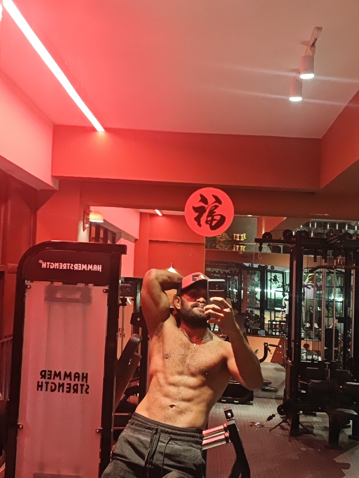 An adult male is captured in a mirror selfie within a gym located in the Chongqing High-tech Zone, China. The individual is shirtless, displaying a muscular physique, and wears a red baseball cap, dark grey sweatpants, and a necklace with a cross pendant. He holds a black smartphone in his left hand to take the photo, while his right arm is flexed behind his head. The gym interior is illuminated by prominent red ambient lighting from LED strips on the ceiling, complemented by some white spotlights. A large mirror reflects the subject and various gym equipment, including a machine with the reversed "HAMMER STRENGTH" logo visible on the left side of the reflection, and other weight machines and racks on the right. Above the subject's reflection, affixed to a red wall section, is a red circular sign featuring the black Chinese character "福" (Fú).