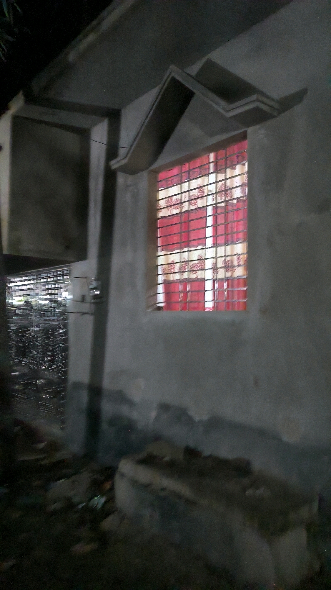 This image depicts the exterior of a building at night in Begumganj Upazila, Bangladesh. The building has a rough concrete finish. A window with a metal grate is visible, covered by red and white patterned curtains. Above the window, there is an angular concrete overhang. To the left of the window, another section of the building is visible, with a large, dark opening that appears to lead into a cluttered interior with rows of objects, possibly shelves or structures. The ground in front of the building is covered in debris and what appears to be rubble, with some indistinct shapes suggesting concrete blocks or construction materials. The lighting is dim, suggesting it is nighttime.