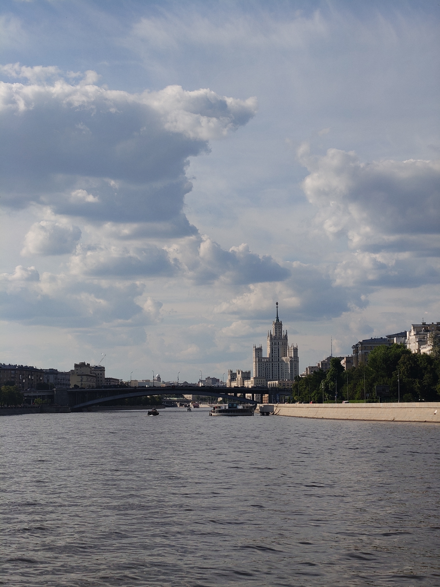 The image displays a panoramic view of a city riverfront on a partly cloudy day. In the foreground, a wide river stretches across the frame, its surface textured with ripples and reflections. Several boats, including at least two small motorboats and a larger passenger vessel, are seen navigating the water.
The midground features a large arch bridge crossing the river, with additional bridges and city infrastructure visible in the distance. The riverbanks are lined with a mix of buildings and trees. On the right, a long concrete embankment runs along the water.
Dominating the skyline in the background is a prominent, multi-tiered Stalinist skyscraper, topped with a spire, characteristic of the "Seven Sisters" architecture in Moscow, specifically resembling the Kotelnicheskaya Embankment Building. Other urban buildings, ranging from apartment blocks to commercial structures, extend along both sides of the river. The sky is filled with large, fluffy clouds, varying in shade from bright white to light grey, with patches of blue sky visible, indicating a partly overcast but bright day.
The lighting suggests it is late afternoon or early evening, with soft, diffused sunlight. No individuals are clearly visible, but the presence of boats and city structures implies active urban life. There is no visible text within the image. The scene conveys a bustling cityscape, strongly hinting at Moscow.