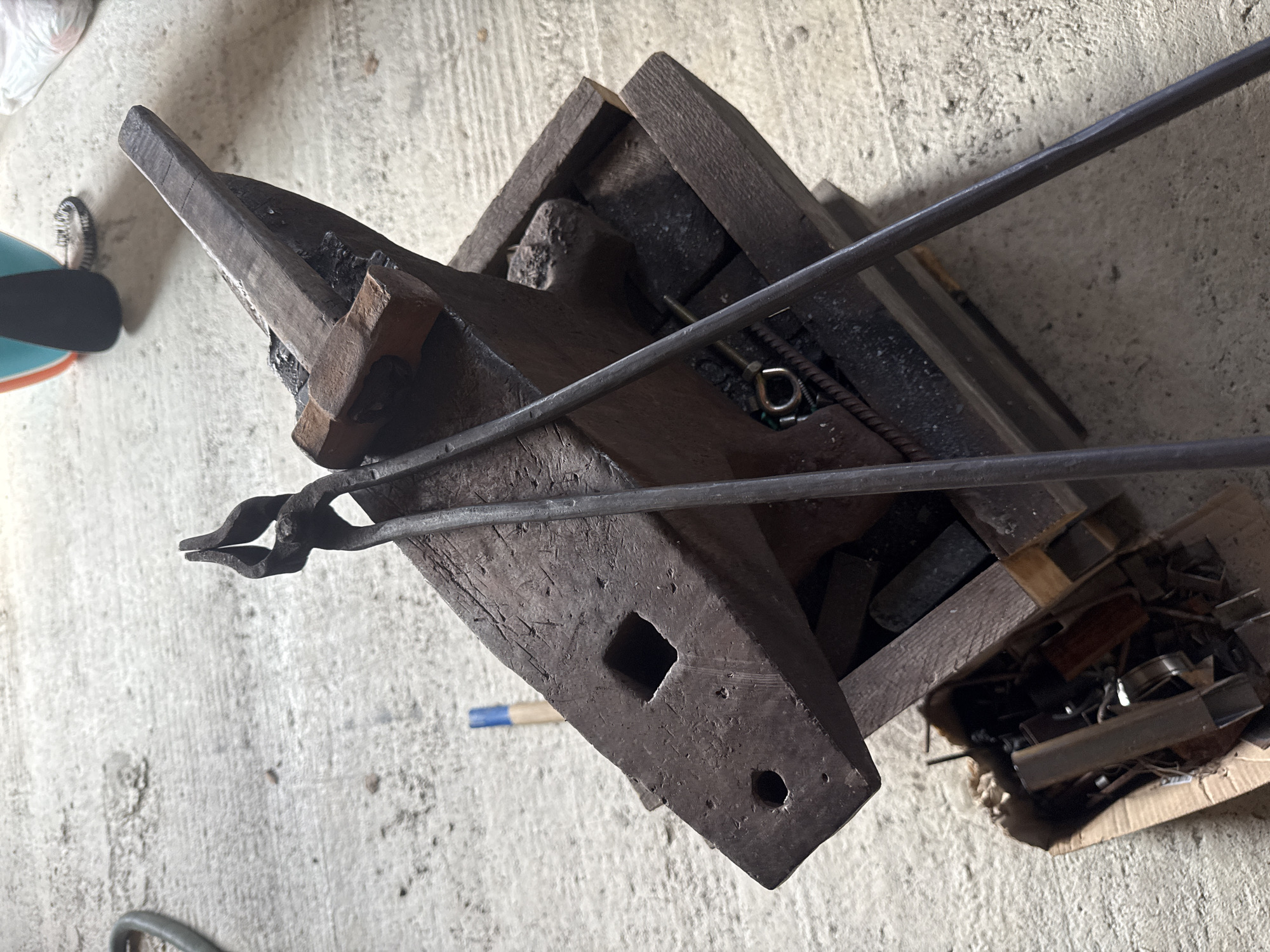 The image shows a close-up, overhead view of a blacksmith's anvil and tools on a concrete floor. The anvil is a heavy, dark metal object with a flat top surface and a horn. It is resting on a wooden stand. A pair of metal tongs is laid across the top of the anvil, with their jaws open. A small wooden-handled hammer with a metal head is also on the anvil. To the lower left of the anvil, a cardboard box is filled with various metal objects, possibly scrap metal or unfinished pieces. A blue-tipped tool is visible on the concrete floor behind the anvil. The environment appears to be an outdoor or workshop setting, with natural light illuminating the scene. No people are present. The scene is located in the city of Krutaya, Russia.