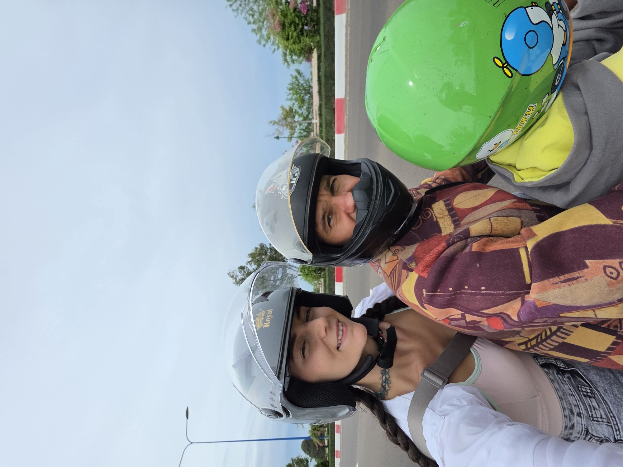 Two people are riding a scooter in Phu Thuy Ward, Vietnam. The foreground shows a woman wearing a white shirt, denim jeans, and a gray motorcycle helmet. A long, dark braid hangs down her back. Behind her, a person wearing a multi-colored jacket and a black motorcycle helmet can be seen. To the right of that person, a green helmet with cartoon graphics is visible. They are on a road with a red and white curb. Behind them is a blue sky and some green trees. Streetlights are visible along the road.