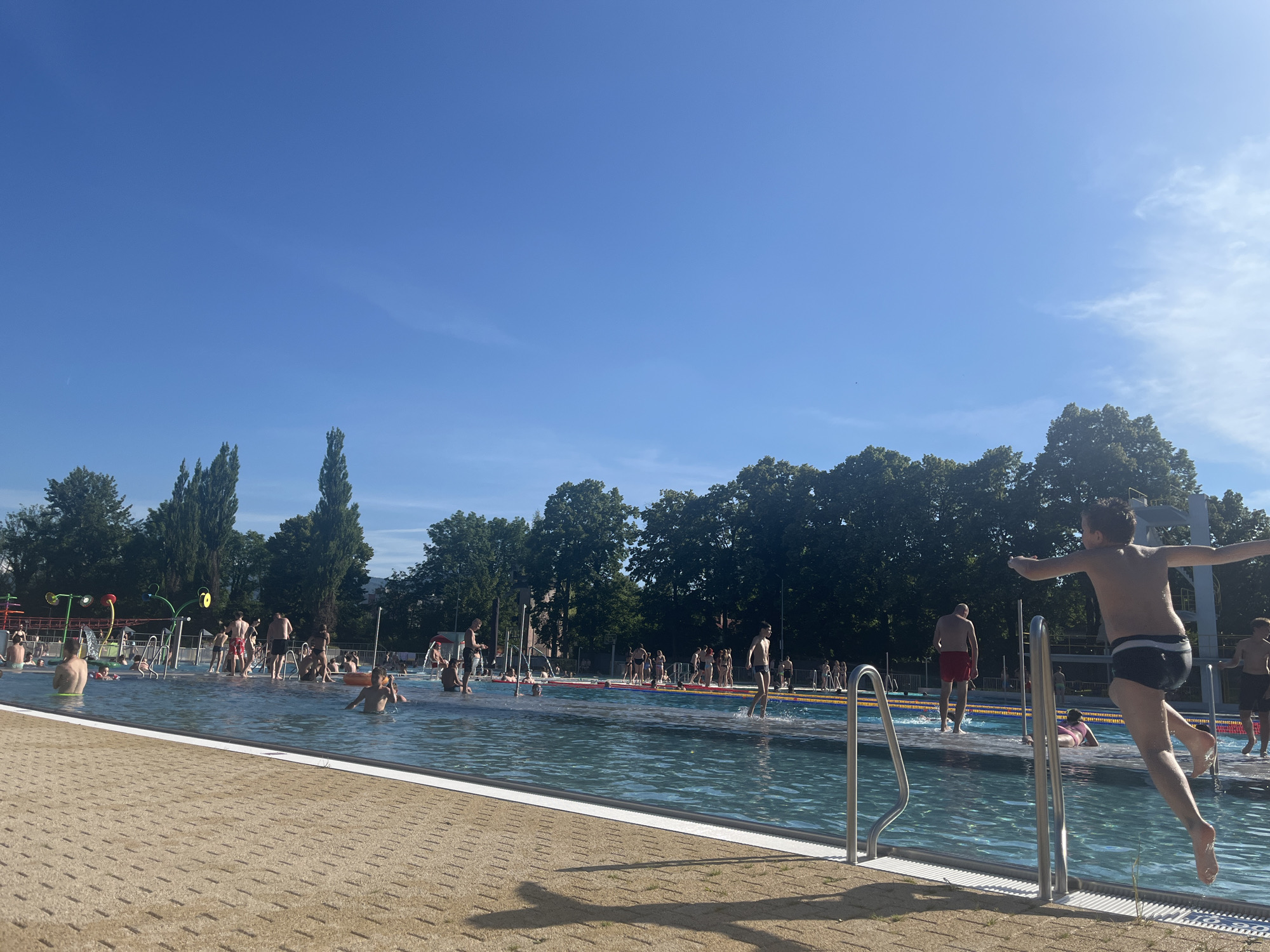 The image depicts a lively outdoor swimming pool on a bright, sunny day with a clear blue sky. In the foreground, a young boy is captured mid-air as he jumps into the pool, arms outstretched and legs bent. He is wearing black swim trunks with a white stripe. The pool itself is a vibrant blue, with several people visible enjoying the water, including adults and children. Some are standing or walking in the shallow end, while others are swimming or playing. In the background, there is a line of lush green trees, providing a natural backdrop to the urban setting of the pool. A diving platform is visible to the right of the jumping boy. The poolside area is paved with beige tiles and features metal railings.