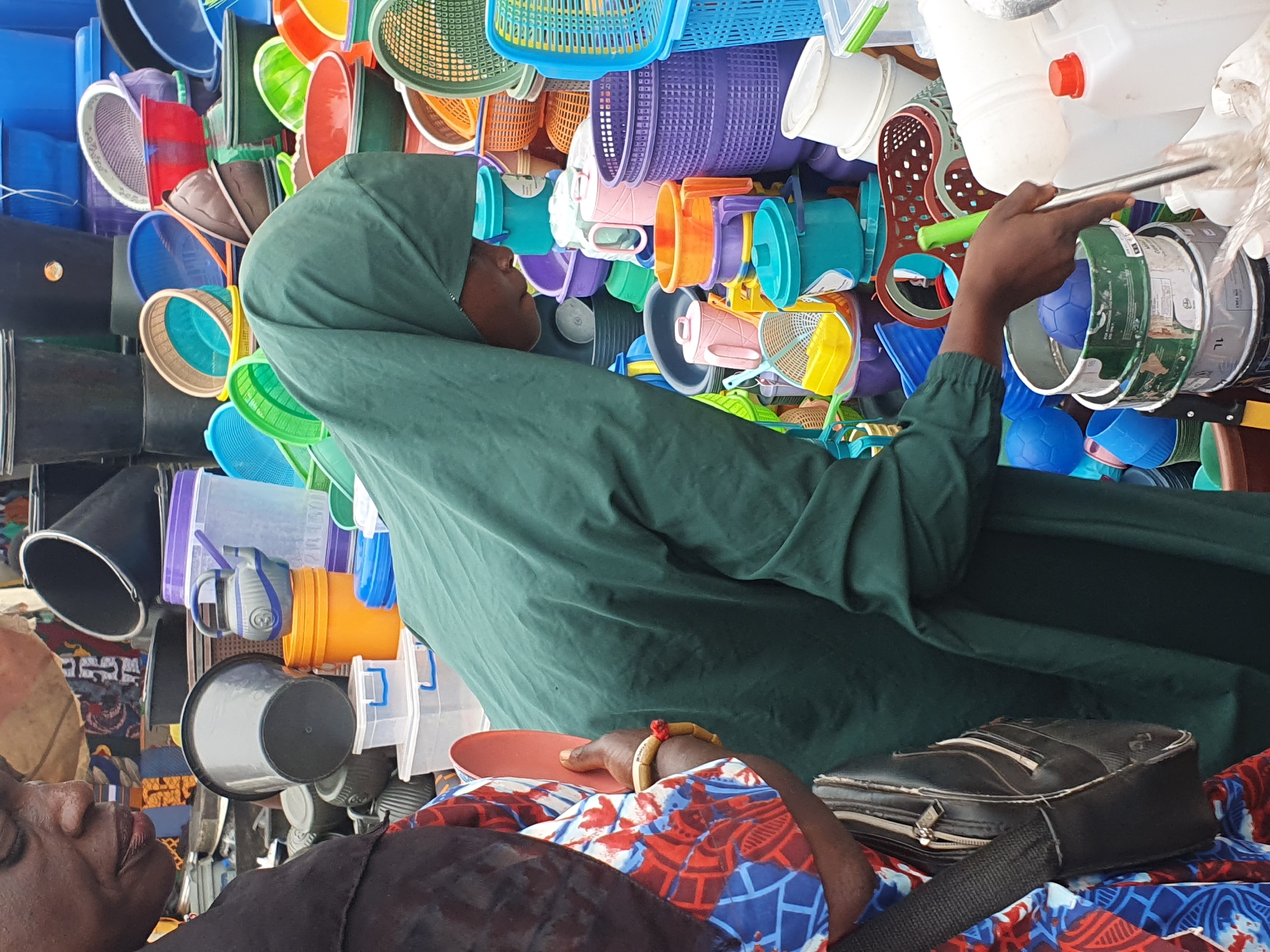 The media file shows a close-up, low-angle, tilted view of a woman wearing a dark green hijab and a patterned fabric. She appears to be in a market stall filled with an assortment of colorful plastic items, including buckets, basins, and containers. The sheer volume and variety of plastic goods suggest a retail setting, possibly a market in Lokoja, Nigeria, as hinted by the location context.
The woman's face is partially visible, and she is looking upwards and to her right, with her eyes closed, suggesting she might be resting or sleeping. Her right hand, adorned with a bracelet, is resting on a red dish, and her left hand is out of frame. Beside her, a black bag with zippers is visible, also resting on the patterned fabric. The overall impression is one of a vendor taking a break amidst her wares in a bustling marketplace. The lighting suggests it could be daytime, with no specific weather cues visible. The arrangement of the plastic items is dense, creating a visually crowded background, predominantly featuring shades of blue, green, red, orange, and purple. The tilt of the image adds a dynamic perspective to the scene.