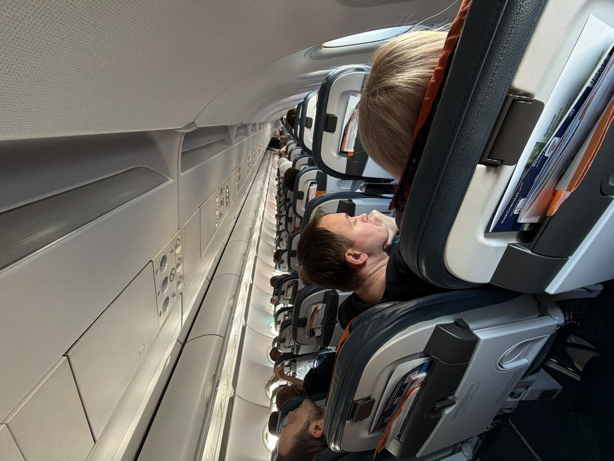 The image depicts the interior of an airplane cabin, viewed from a seated perspective. Rows of airline seats with blue upholstery and grey headrests extend into the distance, some occupied by passengers. Overhead compartments and air vents are visible along the ceiling. The lighting appears to be a mix of natural light from the windows, which are not directly visible but implied by the illumination, and interior cabin lights. The scene is forensic and matter-of-fact, focusing on the environment and the people within it without any specific narrative. The location is specified as a commercial airplane, likely in flight or preparing for departure/arrival, and could be anywhere, including en route to or from the city of Khimki, Russia.