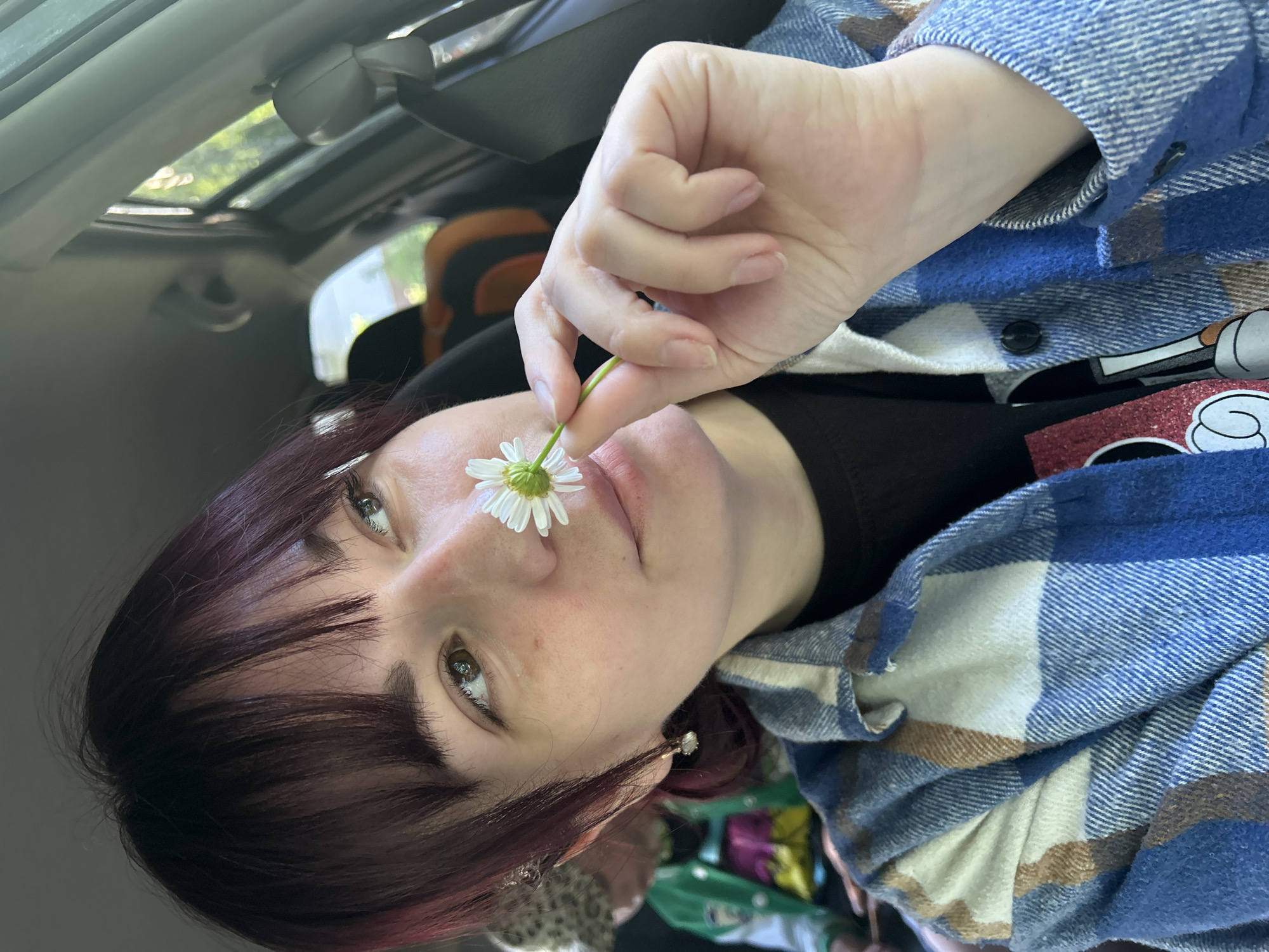 A young woman with dark hair is sitting in the passenger seat of a car, holding a daisy up to her nose. She is wearing a blue and white plaid shirt over a black t-shirt. In the background, another person is visible, wearing a patterned hat and a green jacket. The interior of the car is dimly lit, with sunlight filtering in through the windows. The image appears to have been taken during the daytime, likely in a suburban or rural setting. The specific location in Chaikovsky, Russia, cannot be determined from the visual information provided in the image.