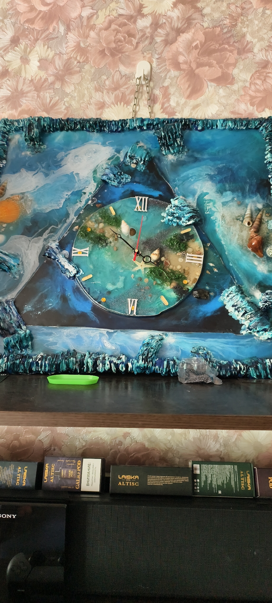 The image captures an indoor scene, primarily featuring a decorative wall clock mounted on a floral wallpapered wall, with various items arranged on a shelf below.
The central subject is a triangular-framed wall clock, crafted in an artistic style resembling an ocean or underwater scene using epoxy resin. The clock face itself is circular, displaying Roman numerals (XII, VIII, VI, XI are visible) and features miniature shells, a starfish, and plant-like elements embedded in a teal, sandy, and dark blue background. The hour and minute hands are black, and the second hand is red. The broader triangular frame surrounding the clock face continues the aquatic theme with textured, wave-like, and coral-like resin formations in shades of blue and white, incorporating larger shells. The clock is suspended by a silver chain from a white adhesive hook on the wall.
Below the clock, on a dark shelf, a small, bright green, oval-shaped dish or tray is placed. To its right, a translucent, crystalline decorative object is also visible. Further down, at the bottom of the frame, a black electronic device, possibly a soundbar or speaker system, with a partial "SONY" logo, is visible. On top of this device, several rectangular product boxes are arranged. Text on these boxes includes "LAISKA", "ALTISC", and "GALIO POD". One box prominently displays a warning in Russian: "ВНИМАНИЕ: Никотин вызывает зависимость" (ATTENTION: Nicotine causes addiction).