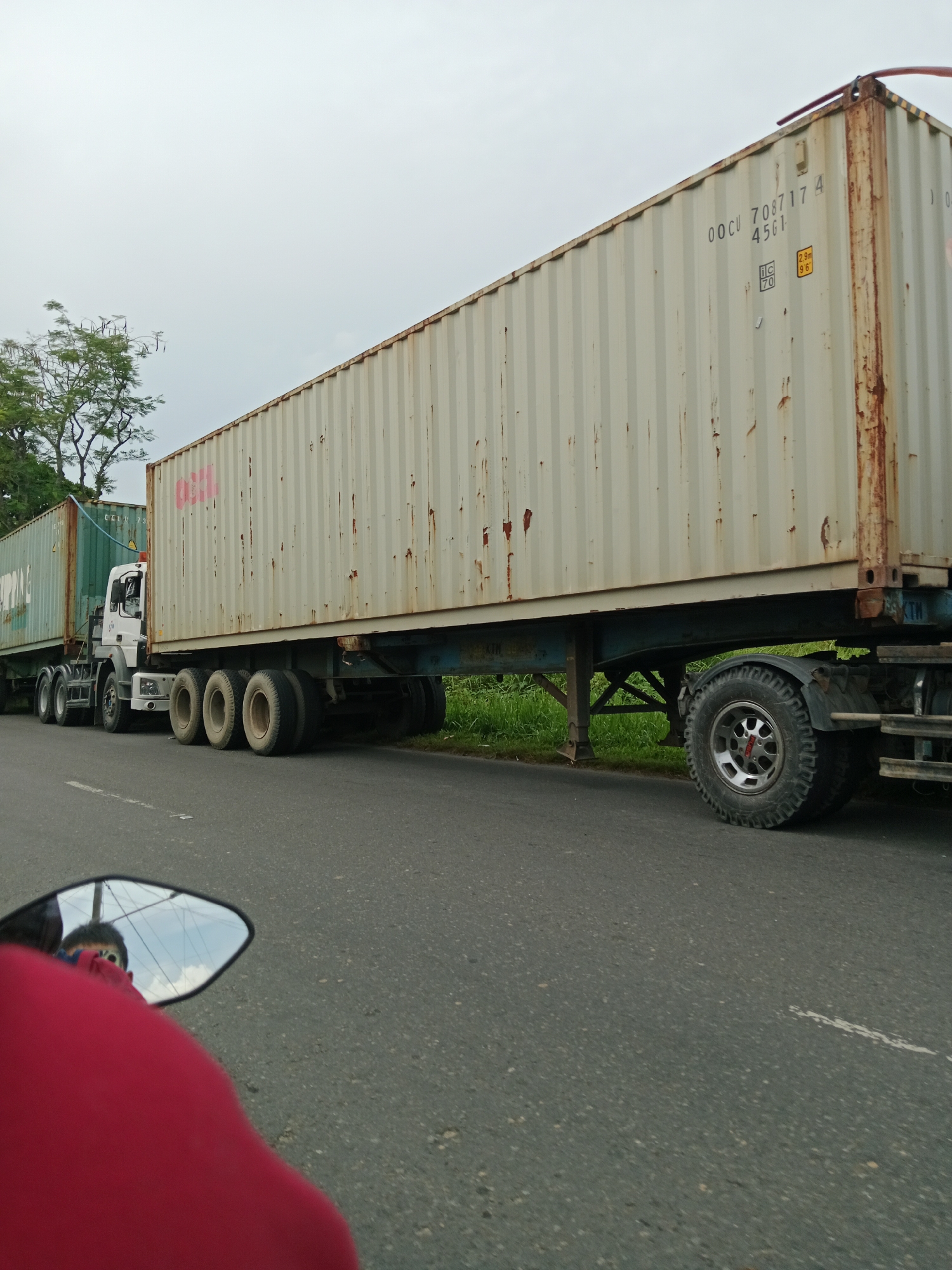 The image, taken from a motorcycle in the City of Binjai, Indonesia, depicts a street scene dominated by two large, multi-axle trucks transporting shipping containers. The truck in the foreground is white with a light-colored, ribbed shipping container. The container has visible rust streaks and markings, including the code "00CU 708717 4 45G1" and yellow and white hazard symbols. A second, dark green shipping container is visible behind the white one, mounted on a separate trailer. The trucks are on a paved road with a white dashed line. To the left of the trucks, there are trees. The sky is overcast. In the foreground, a motorcycle's side mirror reflects the rider, wearing a red shirt, and the sky. The overall environment suggests an industrial or port area, or a major transport route within the city.