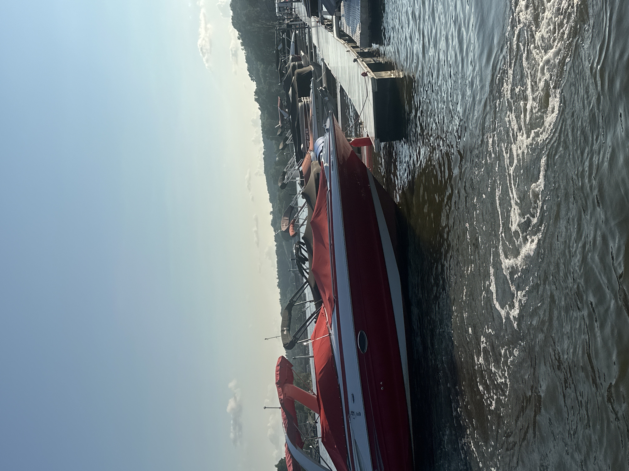 A fleet of boats is moored at a dock on a body of water under a clear sky with scattered clouds. The boats, mostly covered with red and black tarps, are lined up along a wooden pier. One red and white speedboat is prominently featured in the foreground. The water exhibits a slight ripple effect, with some areas of foamy churn. Lush green trees and hills form the background, suggesting a natural setting. The scene is likely in Algonquin, Illinois, a city known for its picturesque waterfront and recreational opportunities.