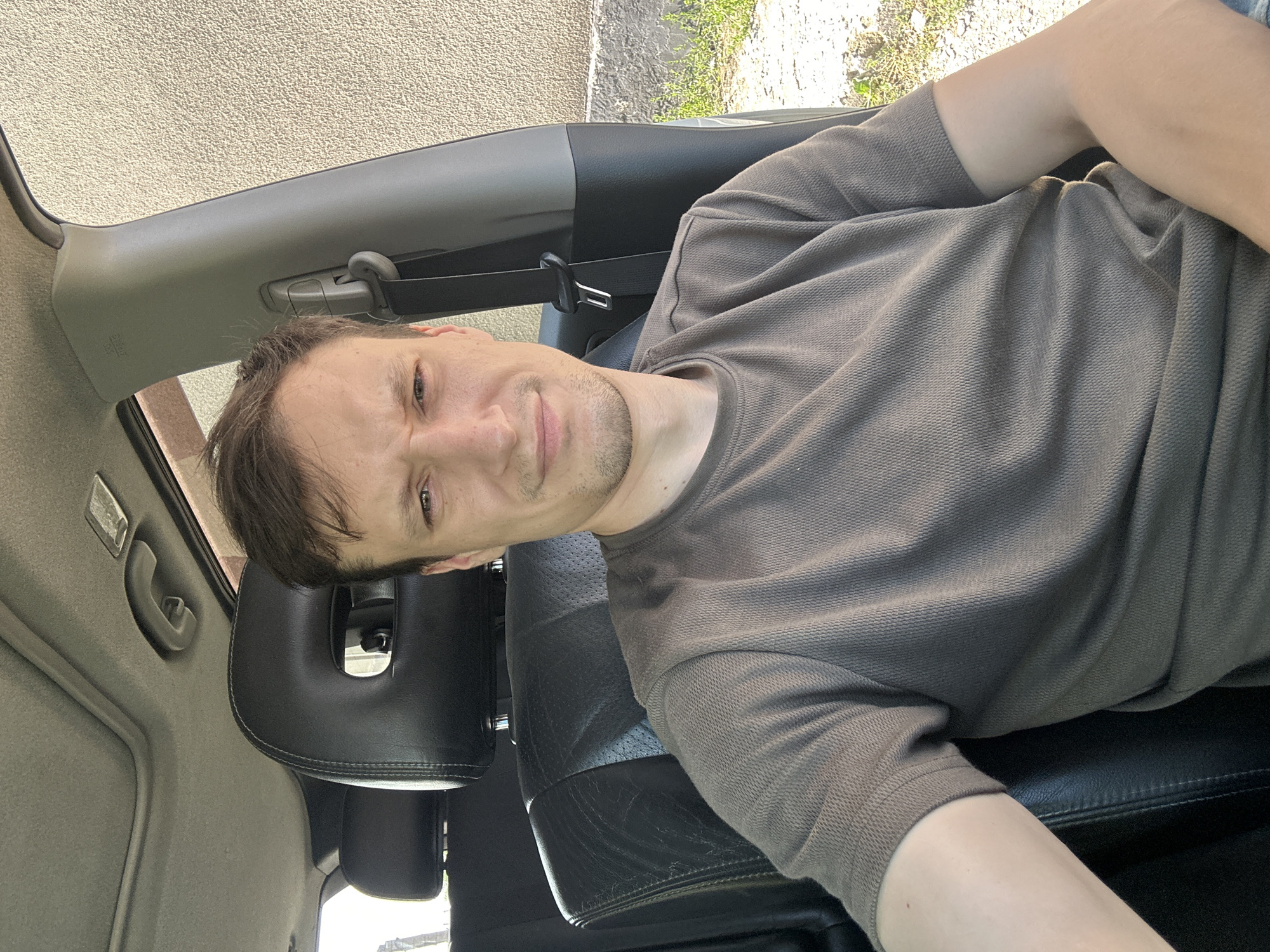 A man with brown hair is sitting in the driver's seat of a car. He is wearing a brown t-shirt. The car has black leather seats and a sunroof. The exterior of the car appears to be parked on a street with a textured wall in the background. The location is Shatava, Ukraine.