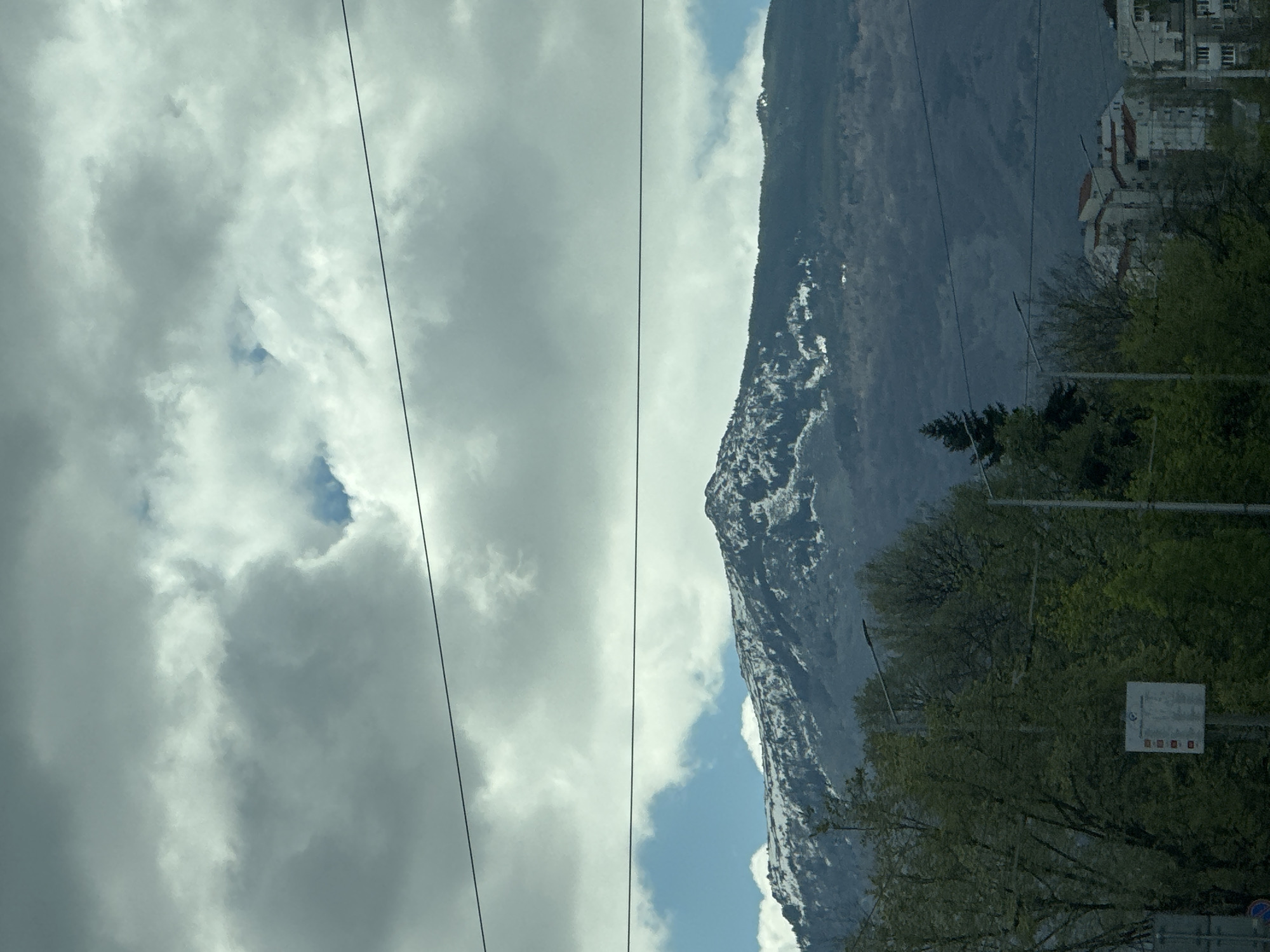 The image shows a view in Sofia, Bulgaria, featuring a mountain partially covered with snow on its slope, and a cloudy sky with visible power lines. The lower right portion of the image includes trees, buildings, and a traffic sign, indicating an urban area.