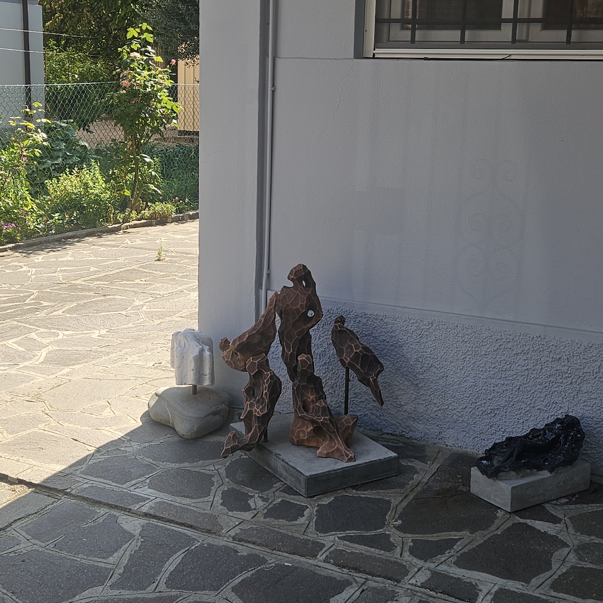 The image depicts an outdoor scene in Modena, Italy, featuring several sculptures placed on a flagstone patio. The foreground is dominated by the patio, made of irregularly shaped, grey flagstones with visible grout lines. To the right of the patio, a white textured wall forms the background, with a rougher, darker textured lower section. A window with a dark frame and panes is visible high on the wall.
Several sculptures are arranged on the patio. On the far left, a small, white, abstract marble sculpture rests on a rounded grey stone. Next to it, and more centrally, is a taller, dark brown abstract sculpture composed of interconnected organic shapes, with a small circular element embedded in its center. To the right of this, a smaller, dark brown abstract sculpture, resembling a bird or a bird-like figure, is mounted on a thin black rod. Further to the right, a small, dark, amorphous sculpture sits on a rectangular grey base.
In the background, a chain-link fence encloses a garden area with lush green foliage and some pink flowers, suggesting a sunny day. The lighting indicates the sun is casting shadows across the patio, with a prominent shadow falling from the left. The overall impression is one of an artistic display set within an outdoor, possibly residential or gallery space.