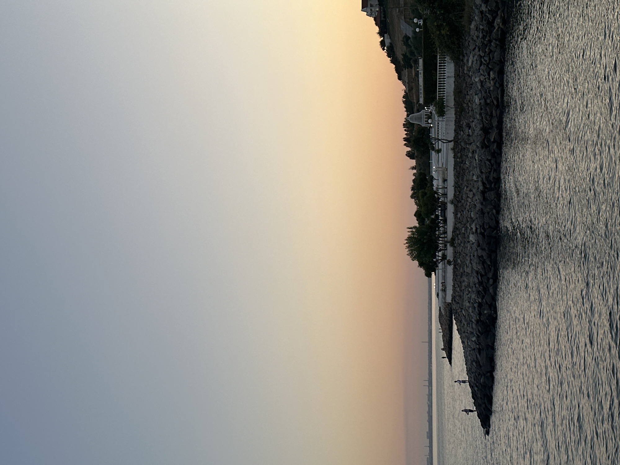 The image depicts a scenic waterfront view in Odesa, Ukraine, during what appears to be sunset or twilight. The sky transitions from a clear pale blue at the top to soft orange and yellow hues closer to the horizon.
In the foreground, a body of water with gentle ripples occupies the lower portion of the frame. Along the mid-ground, a rocky shoreline juts out into the water. On this rocky expanse, two individuals are visible, each engaged in fishing. They are standing, holding fishing rods, facing the water.
Further along the shoreline, a white-fenced promenade or walkway is visible, lined with trees and some architectural structures, including a white gazebo-like building. A hillside with vegetation and a few houses can be seen rising behind the promenade. In the distance, across the water, a faint outline of industrial structures or a cityscape is discernible on the horizon. The overall atmosphere is tranquil and serene.