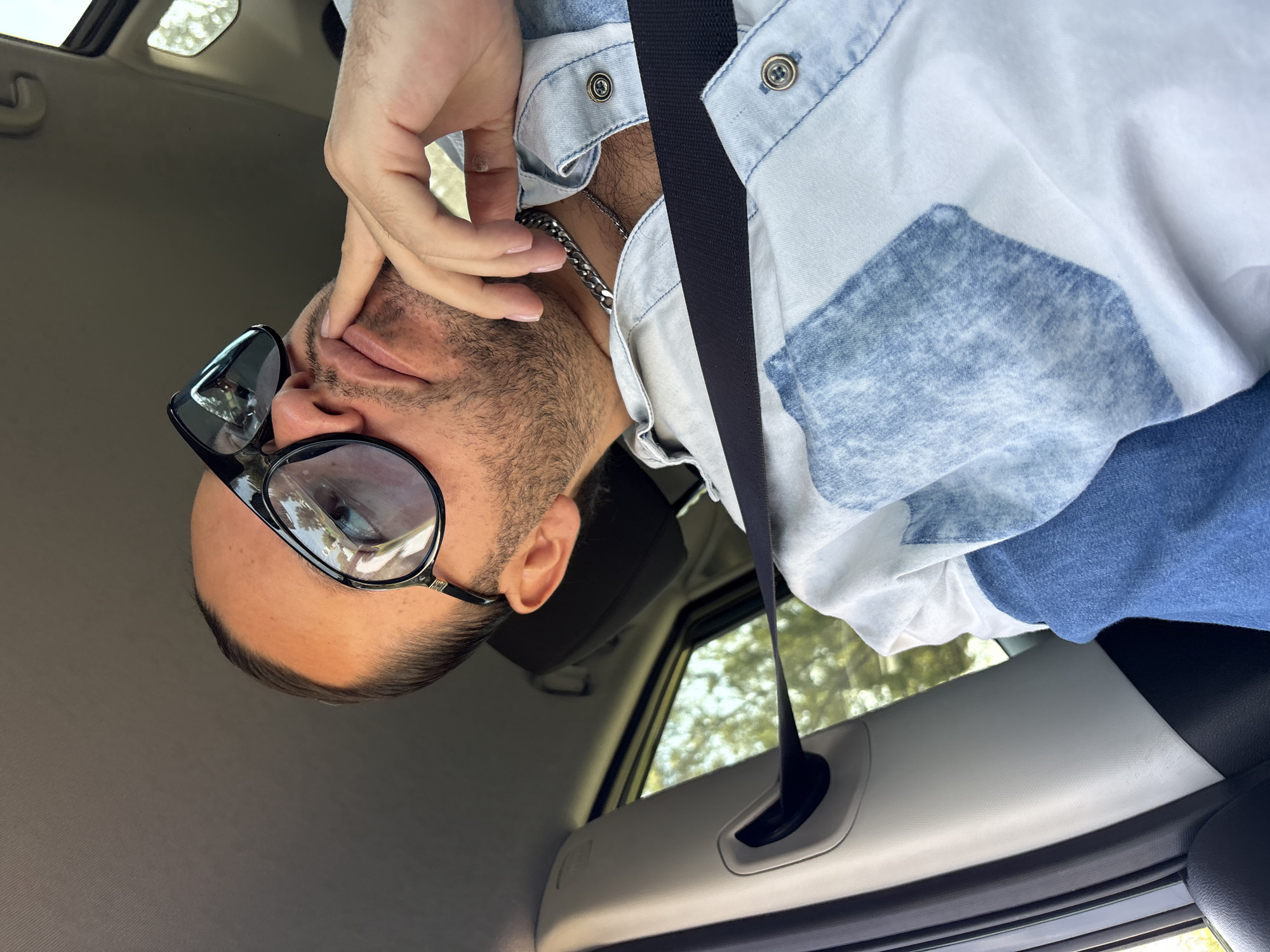 An overhead, rotated shot shows a person, likely male, seated inside a car during what appears to be daytime. The subject, wearing a light blue and white denim-patterned shirt and a metal chain, has their right hand raised to their face, fingers touching their forehead and eyebrow, partially obscuring their eyes. They are also wearing large, dark-framed sunglasses, with reflections visible on the lenses. A short beard and mustache are visible on their face. A black seatbelt crosses their chest and lap, running diagonally across the frame.
The car's interior is visible, featuring light grey and dark grey panels. Through the car window, green trees and bright, diffused light are seen, suggesting an outdoor setting. The angle of the shot, which is rotated 90 degrees clockwise from a typical upright perspective, makes it appear as if the person is looking upwards or the camera was held sideways. The overall setting is consistent with being inside a vehicle in San Andrés Cholula, Mexico. There is no visible text in the image.
