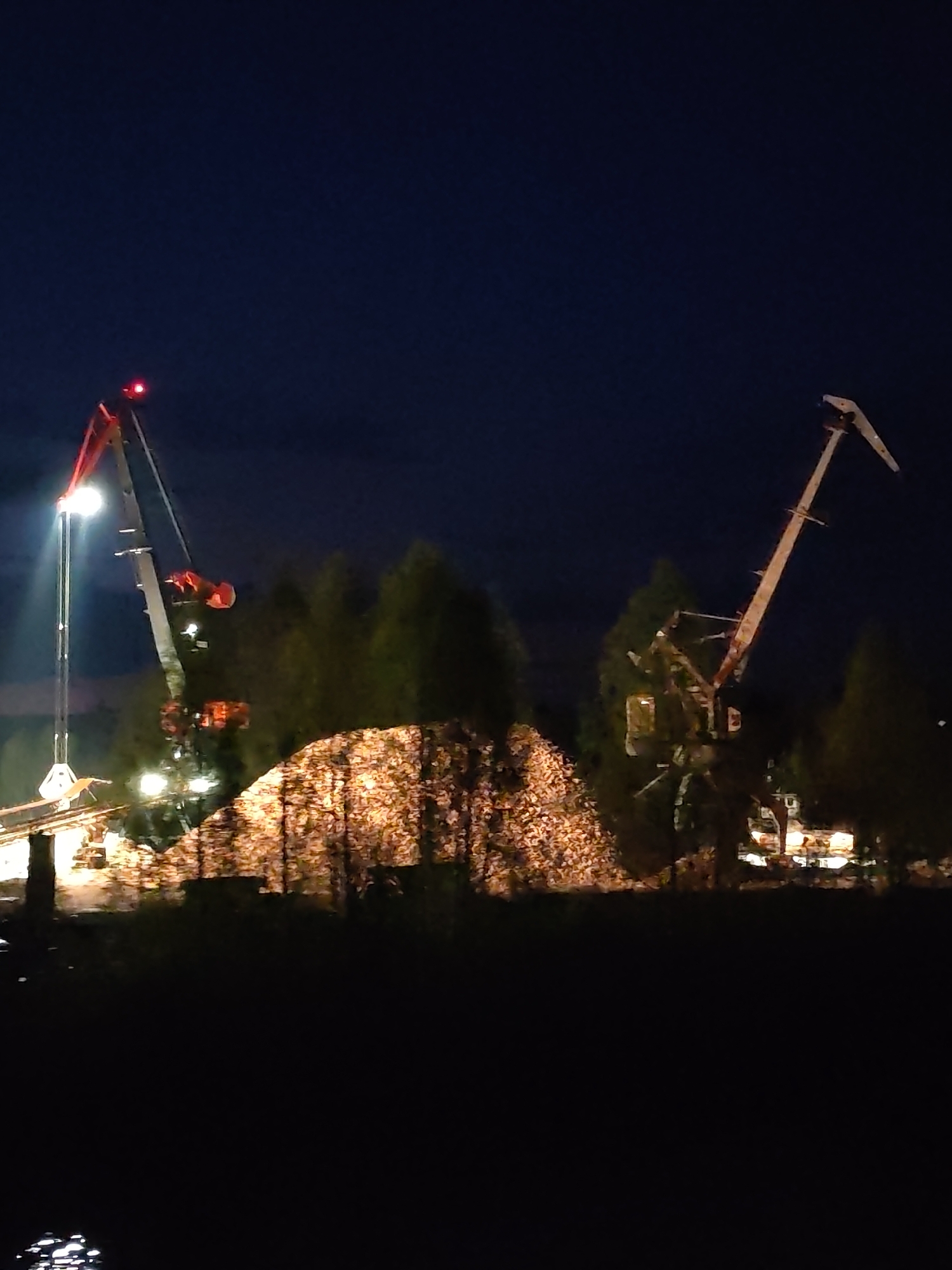 The image depicts an outdoor scene at night, possibly in Александровское, Russia, due to the query. Two cranes are prominent, one on the left illuminated by its own lights, with a red top. A pile of material, possibly wood chips, glows warmly in the center. Trees form a backdrop. The foreground is dark. Reflective patches suggest a body of water in the extreme lower left. The night sky is a deep blue.