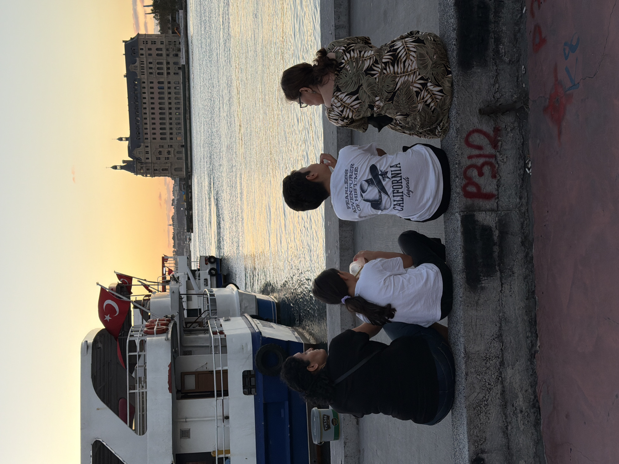 The image depicts a family of four sitting on a concrete ledge overlooking a body of water at sunset. In the background, there are several boats docked, one of which flies a Turkish flag. Across the water, a large, ornate building with multiple stories and twin towers stands prominently, identified as the Haydarpaşa Train Station in Kadıköy, Turkey. The sky is a soft orange and yellow, indicating the sun is setting. The family members, two adults and two children, are seen from behind, facing the water and the historic station. The two children are a girl wearing a white t-shirt and a boy wearing a white t-shirt with graphics. The adults are a woman in a black t-shirt and a woman in a patterned dress. The scene conveys a sense of peaceful observation and shared experience by the waterfront.
