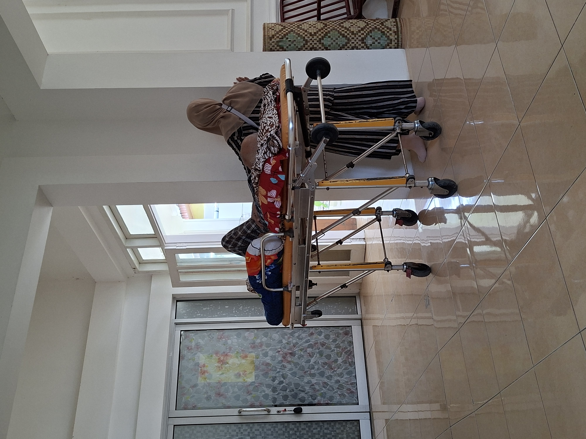 A person in striped clothing is lying on a foldable medical stretcher, viewed from a high angle. The stretcher is indoors on a tiled floor in a building with a large window. The location is Uleereuleung, Indonesia.
