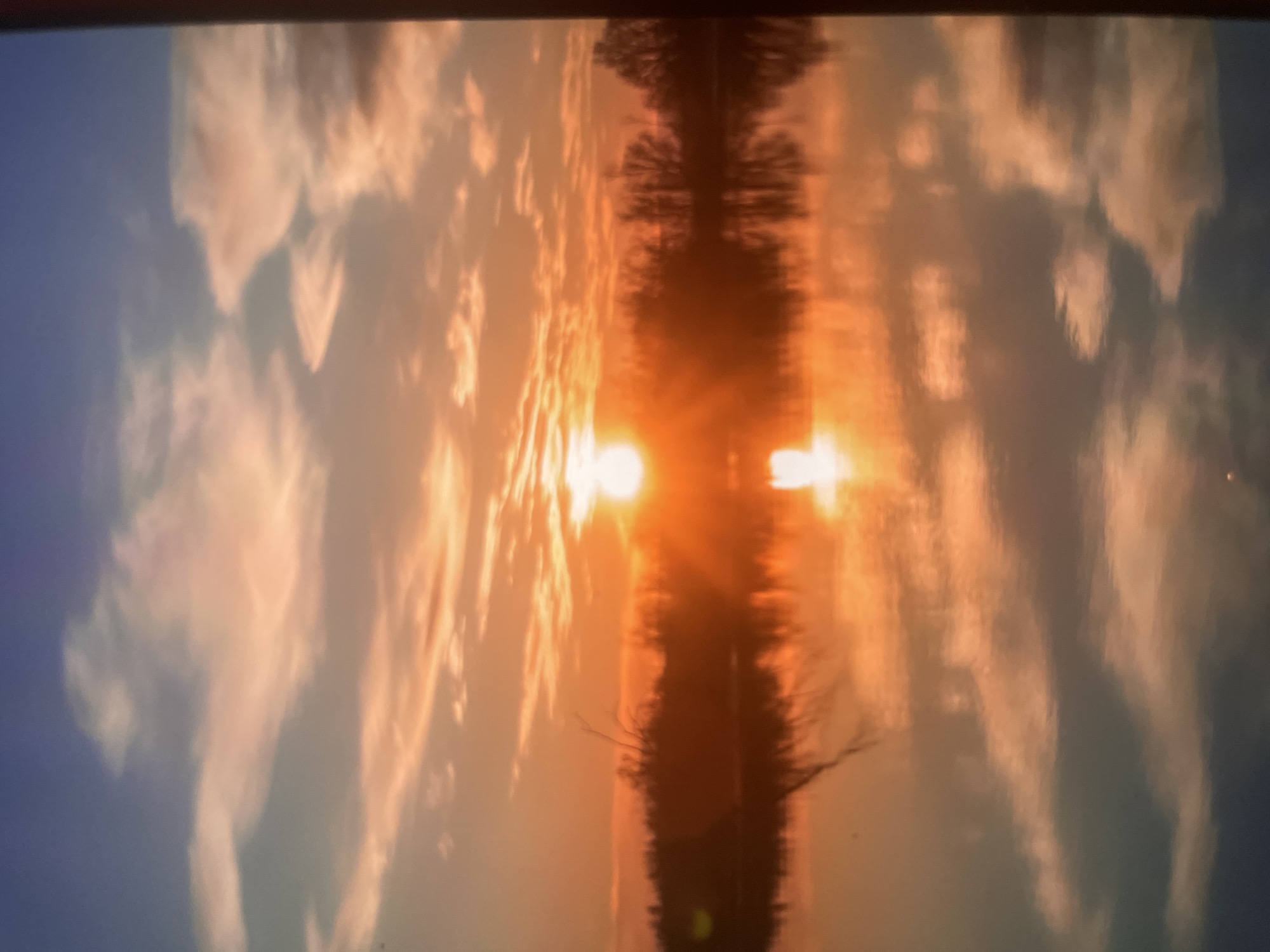 A photograph captures a vibrant sunset over a body of water, reflecting the sky's fiery hues. The sun, a bright orb of light, is positioned near the horizon, casting long rays that illuminate the scattered clouds. The sky transitions from a deep blue at the top to warm oranges and pinks near the horizon. A line of silhouetted trees forms a dark band across the midground, separating the sky from the water. The water's surface is calm, creating a mirror image of the sky and the trees, with ripples subtly distorting the reflection. The scene appears to be a natural landscape, possibly a lake or river, with no human presence or urban structures visible. The image is consistent with a sunset view in Khanty-Mansiysk, Russia, known for its picturesque natural surroundings.