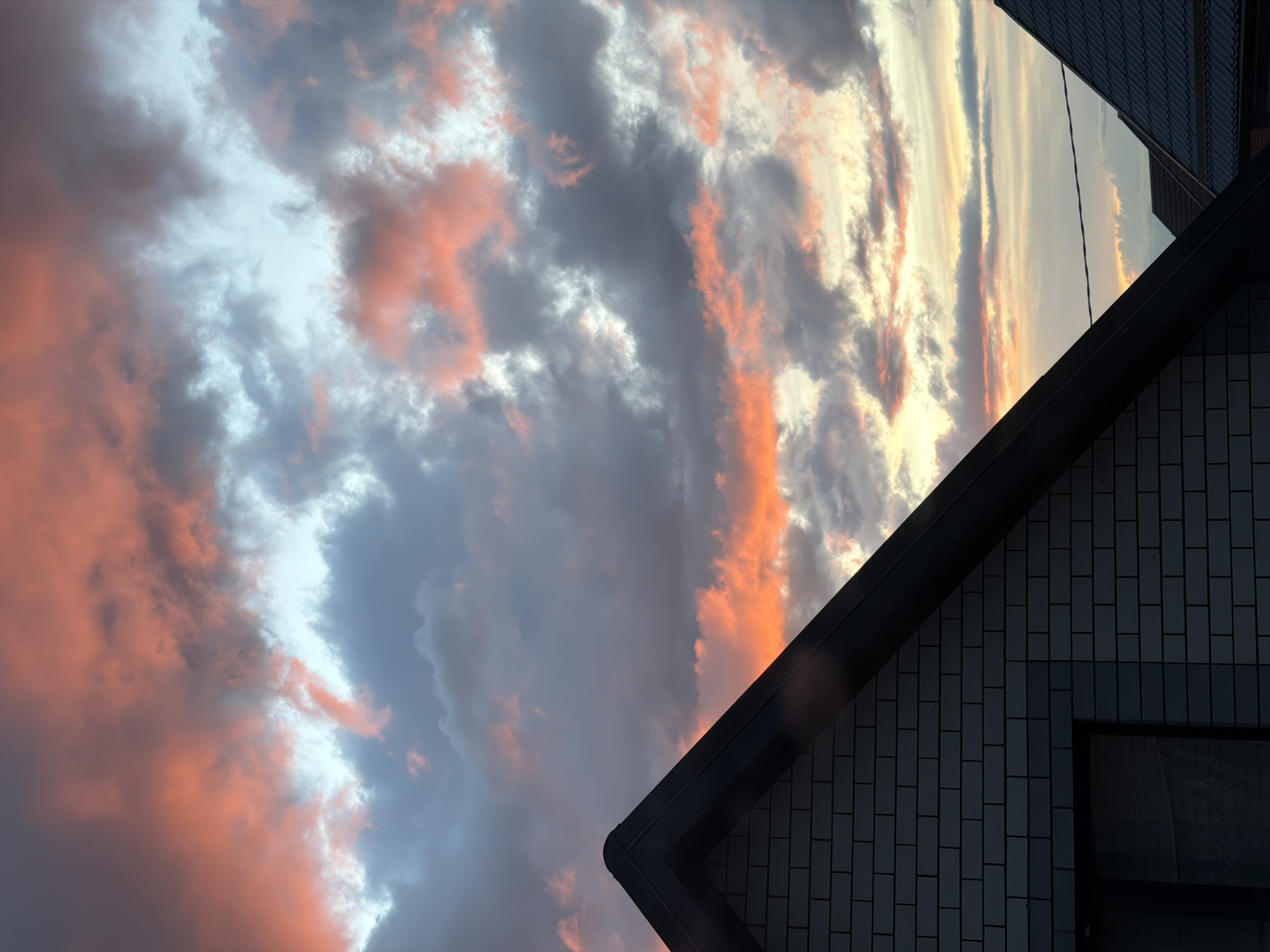 The image captures a dusk sky above a building in what appears to be the Rossiyskiy district of Russia. The sky features dramatic clouds with vivid pink and orange hues intermingling with gray and white. The building's corner, siding (possibly brick), roof, and a window are partially visible in the bottom right corner. The lighting suggests the sun is either setting or has recently set. There are no people visible in the image.