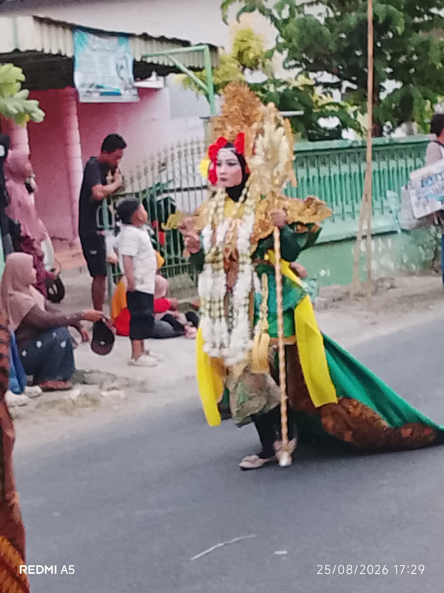 This is a photograph taken in Semanding, Indonesia, on August 25, 2026, at 5:29 PM. The image captures a street scene where a person in elaborate traditional costume is the central focus. The individual is adorned with a gold headdress with red accents, a white and gold layered garment, and a long green cape draped behind them. They are holding a decorative staff.
Around this central figure, other people are present, observing the event or participating in activities related to it. To the left, a woman wearing a light brown hijab and dark pants is seated on the ground, holding a dark cap. Behind her, a man in a black t-shirt is standing. Further back, a young boy in a white striped shirt and dark shorts is looking upwards. Other individuals are visible in the background, some seated on the ground, suggesting a public gathering or performance.
The environment appears to be a street with a paved surface. Buildings with pink and light-colored walls are visible in the background, along with a green metal fence. Lush green trees are also present, contributing to the outdoor setting. The overall impression is of a community event taking place in a semi-urban or village environment in Indonesia.