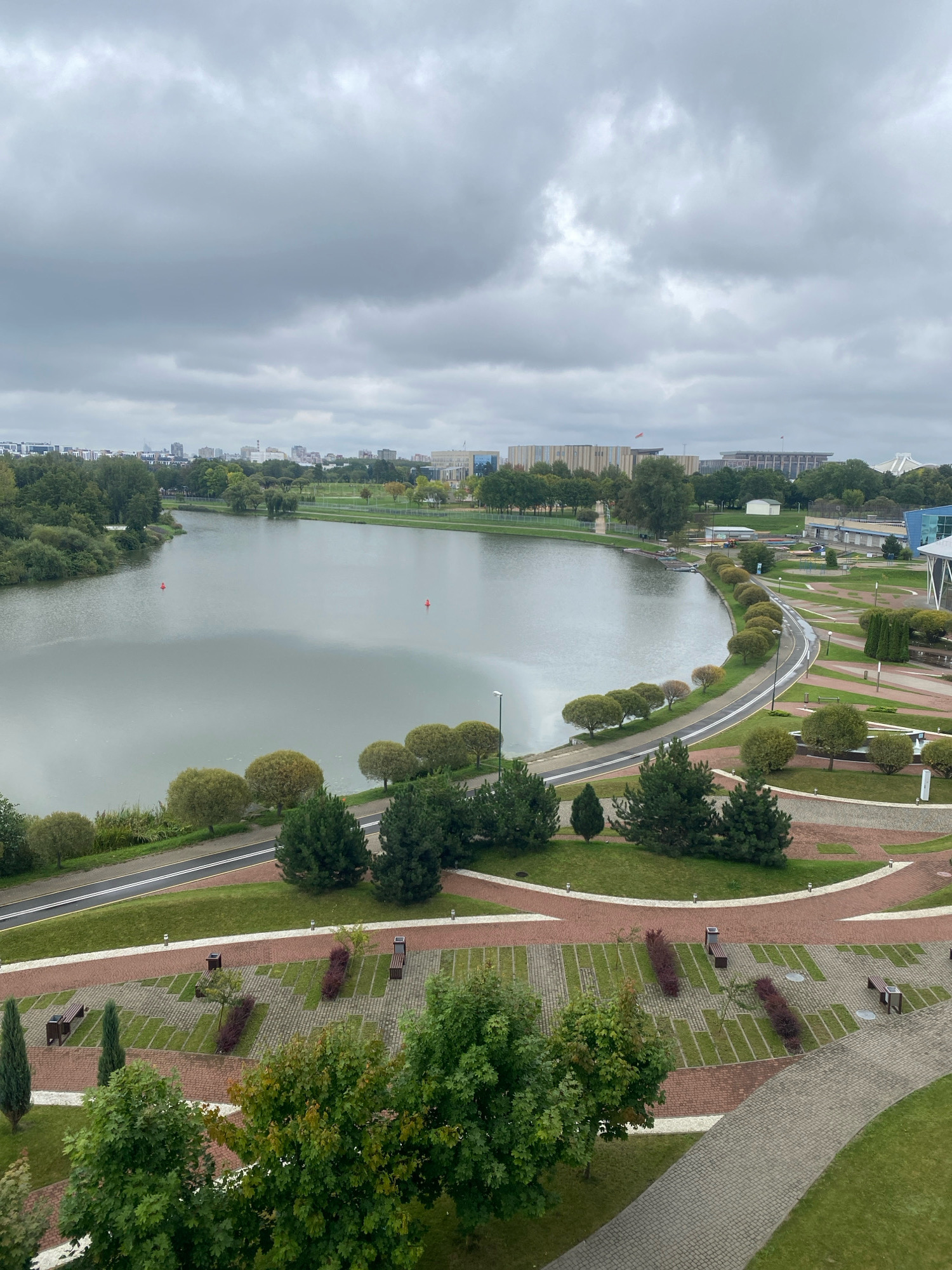 This is an aerial, eye-level, wide shot of a calm lake in a park in Minsk, Belarus, on a cloudy day. The lake is in the center-left of the image and is surrounded by trees and a walking path. In the background, there are buildings and cityscapes, suggesting an urban environment. The foreground features a paved area with benches and manicured greenery. The overall atmosphere is serene and natural, with a clear emphasis on the lake and its surroundings.