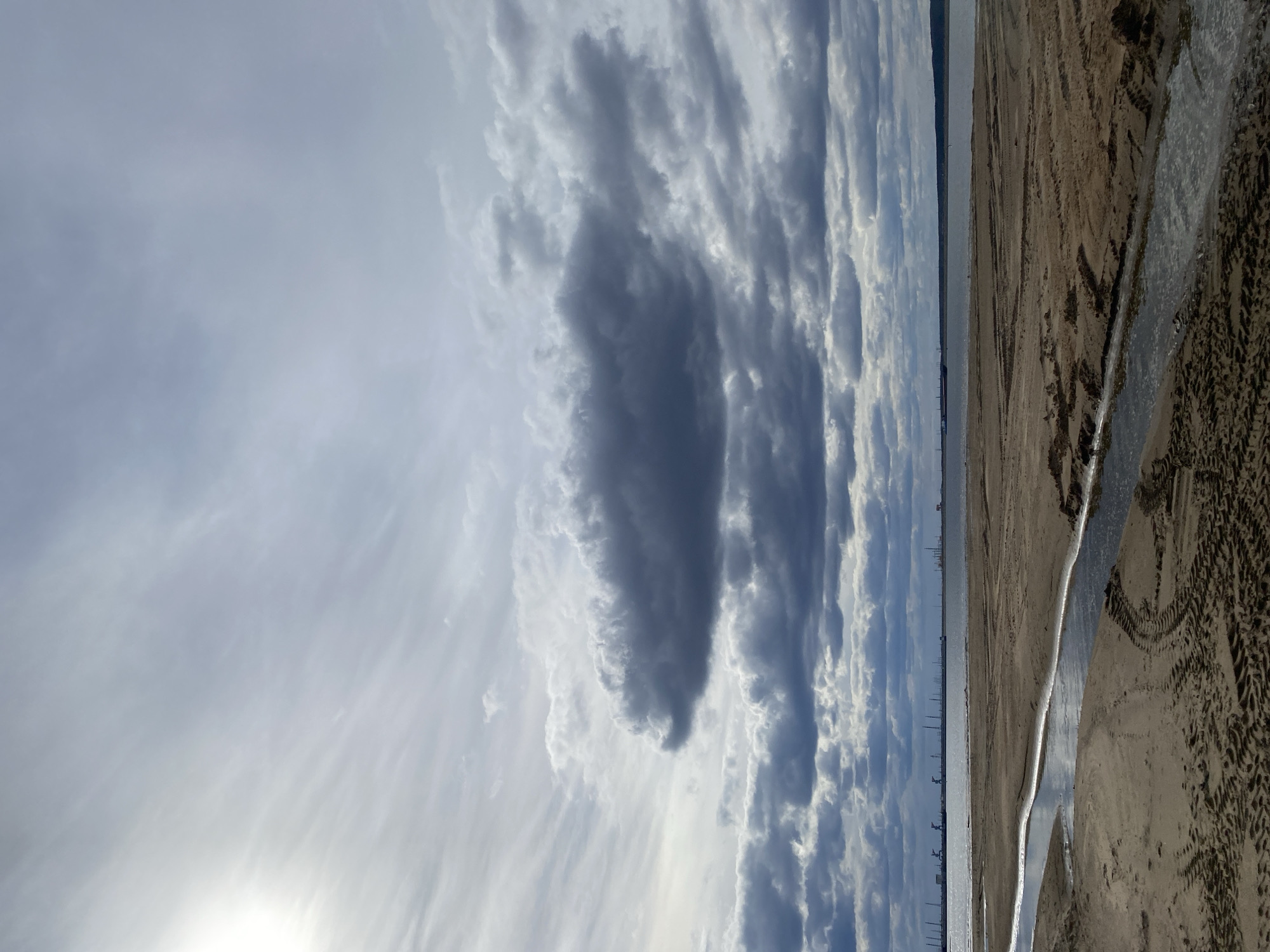 The image displays a panoramic outdoor landscape, with a dramatic sky dominating the upper portion and a sandy shoreline in the foreground.
The sky, occupying roughly two-thirds of the frame, is filled with a dynamic array of clouds. A bright area in the lower left of the sky suggests the position of the sun, casting a strong light. Dark, textured clouds stretch across the upper and middle sections, creating a stark contrast with lighter, more scattered, and wispy cloud formations. The clouds appear layered and show signs of movement or wind, contributing to a sense of vastness.
A faint but distinct horizon line separates the expansive sky from the distant land or water. Along this distant line, subtle vertical structures are visible, possibly indicating buildings, industrial infrastructure, or distant trees on the far shore.
The lower third of the image features a sandy or muddy shoreline. A narrow, winding channel of water cuts through the sand, reflecting the bright sky above. The sand exhibits various textures, including what appear to be tire tracks or other imprints, suggesting some form of past activity, although no people or vehicles are present in the scene.
The time of day is clearly daytime, likely mid-day, given the sun's brightness. The weather is cloudy but illuminated, creating a compelling interplay of light and shadow on the cloud formations. The scene evokes a feeling of openness and tranquility. No specific activity or event is taking place, and there is no visible text. The location, provided as Chaikovsky, Russia, suggests this could be a view along the Kama River or a local reservoir.