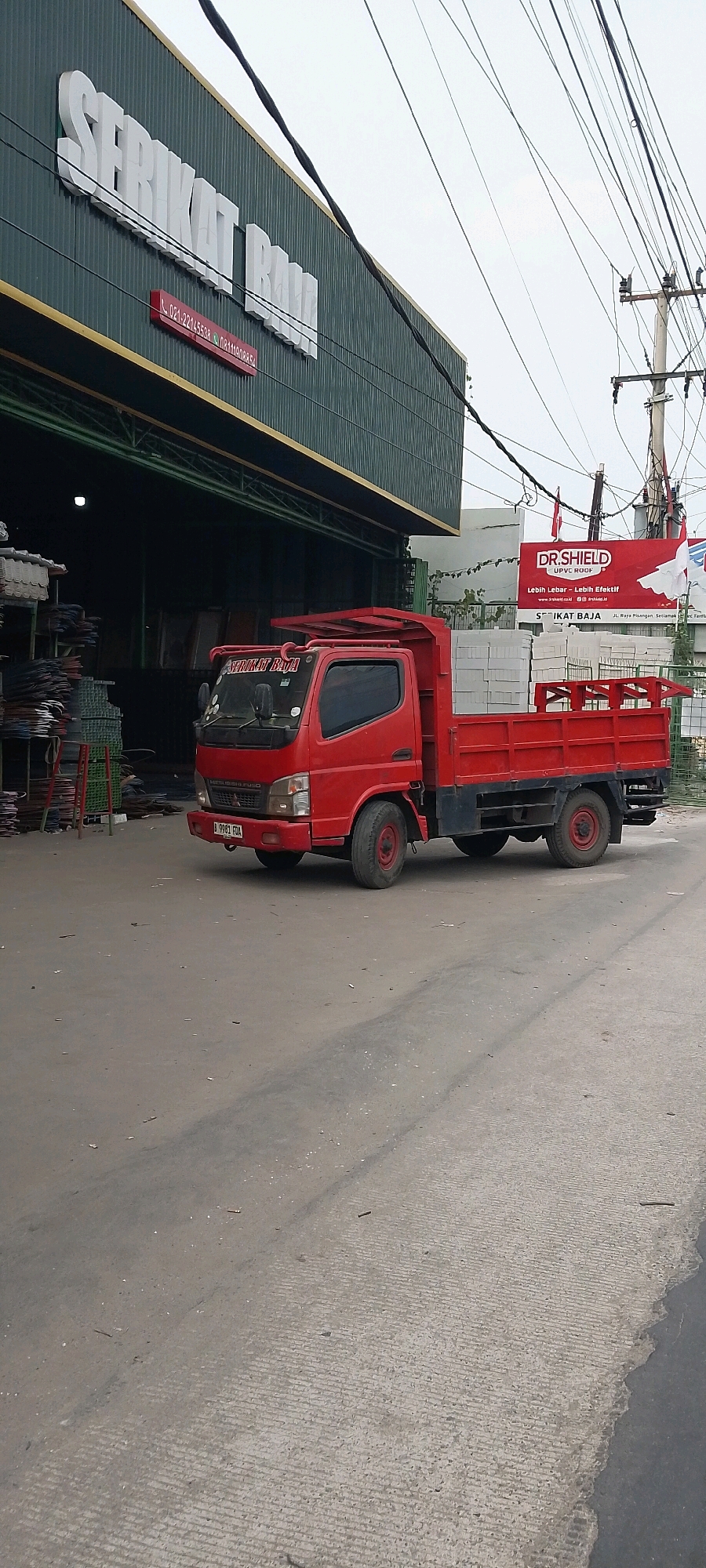 A red Mitsubishi Fuso light truck is parked on a concrete road in front of a building with a dark green corrugated metal facade. The building has large white lettering spelling "SERIKAT BAJA" above a red rectangular sign with white text. The truck has a black bumper, a black chassis, and a red cargo bed filled with white blocks. A license plate reading "I 9981 FDA" is visible on the front of the truck. Stacks of metal and other materials are visible in the open doorway of the building. A red banner with white lettering and a logo that says "DR. SHIELD UPVC ROOF" is attached to a pole to the right of the building. The sky is overcast.