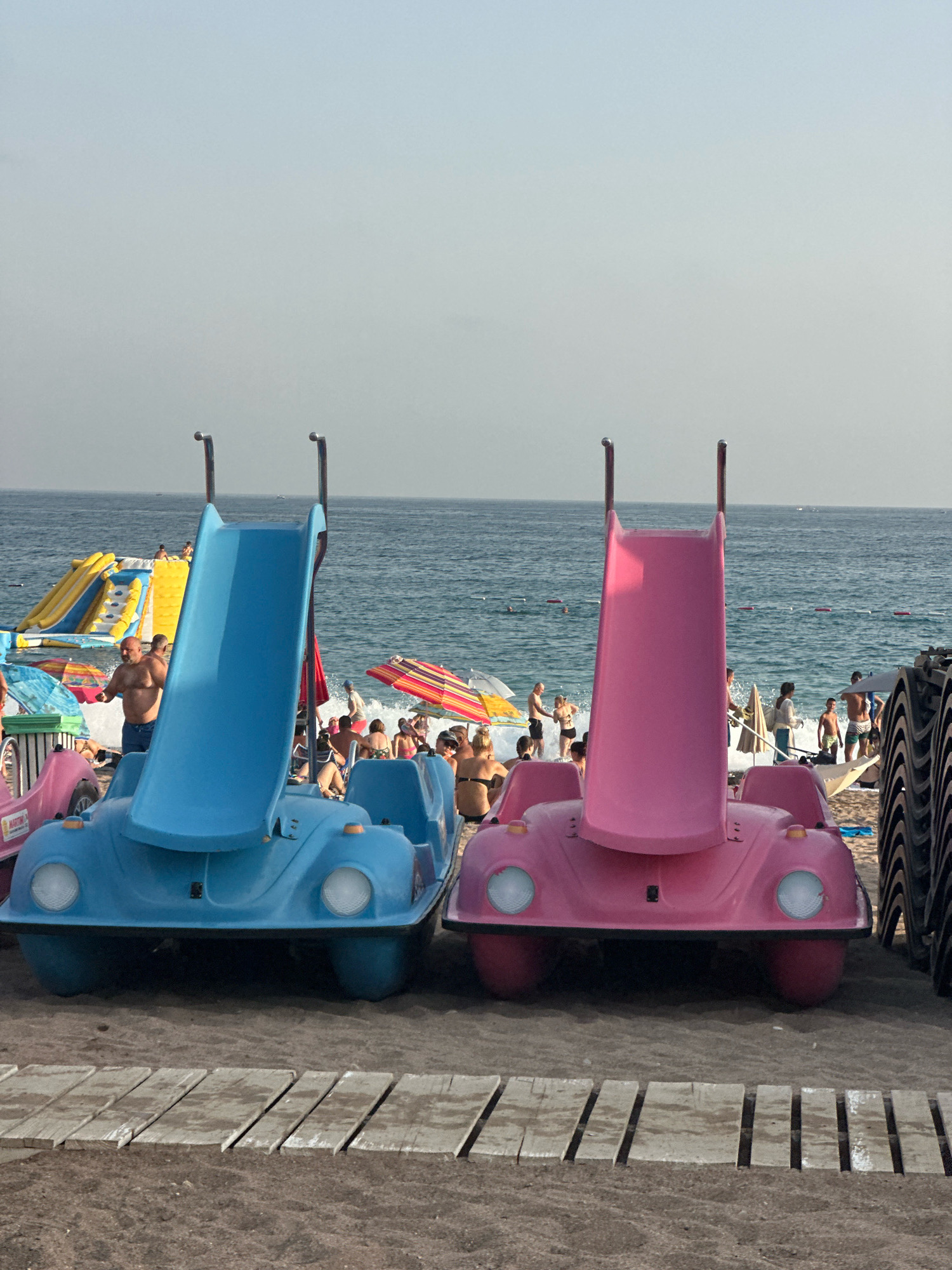In Boreti, Montenegro, two brightly colored pedal boats shaped like vintage cars with slides are parked on a sandy beach. A blue pedal boat with a blue slide is on the left, and a pink pedal boat with a pink slide is on the right. The boats face the viewer and are positioned on a wooden walkway. In the background, the Adriatic Sea is visible with people swimming and relaxing on the beach. Inflatable water slides are set up further down the shore. The sky is overcast.