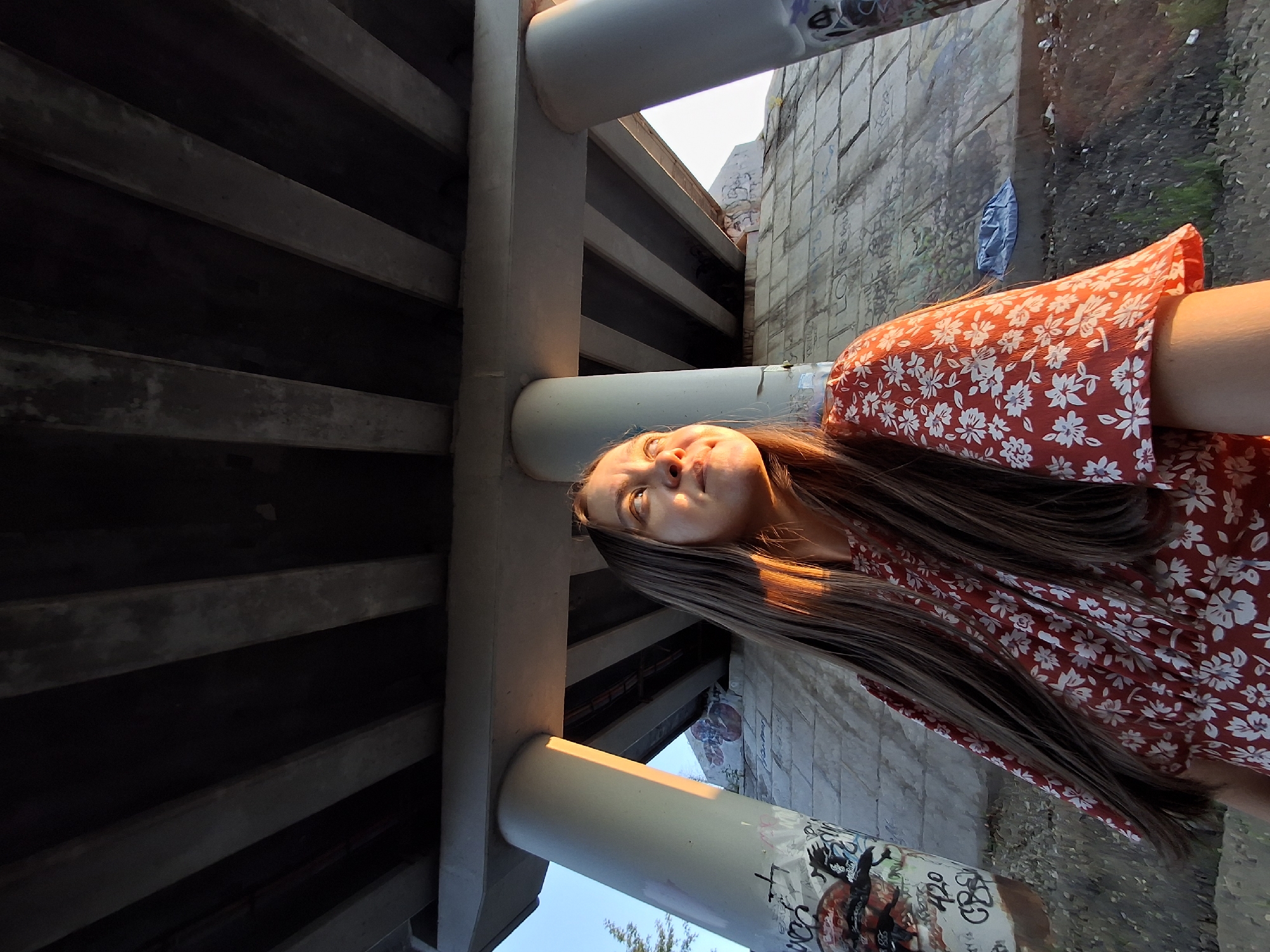 The image shows a young woman standing under a concrete overpass at sunset. The woman has long, light brown hair and is wearing a red dress with a white floral pattern. She is looking up and to her right, with the sun illuminating her face. The overpass is supported by large concrete pillars, and the concrete embankment beneath it is covered in graffiti. The ground is a mix of dirt, gravel, and some sparse vegetation. The sky is clear and blue, with the warm hues of sunset casting long shadows. This location is likely in Chaikovsky, Russia, given the presence of similar infrastructure and the overall environment.