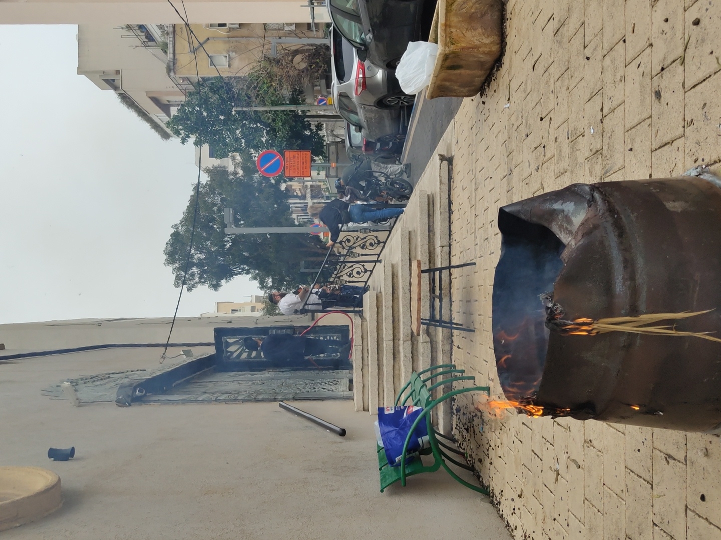 A medium-angle outdoor shot depicts a street scene with a burning barrel, likely occurring in Tel-Aviv, Israel. The barrel, located on a brick-paved sidewalk, emits smoke and flames with dried plant material visibly burning inside. A green metal bench sits near the barrel, with a blue and white bag placed on it.
Beyond the sidewalk, a set of stairs with a black metal railing leads up to a street where pedestrians and a parked car are visible. Street signs can be identified in the distance. The buildings on the left are off-white, while the building on the right is taller with multiple floors. The sky appears overcast.