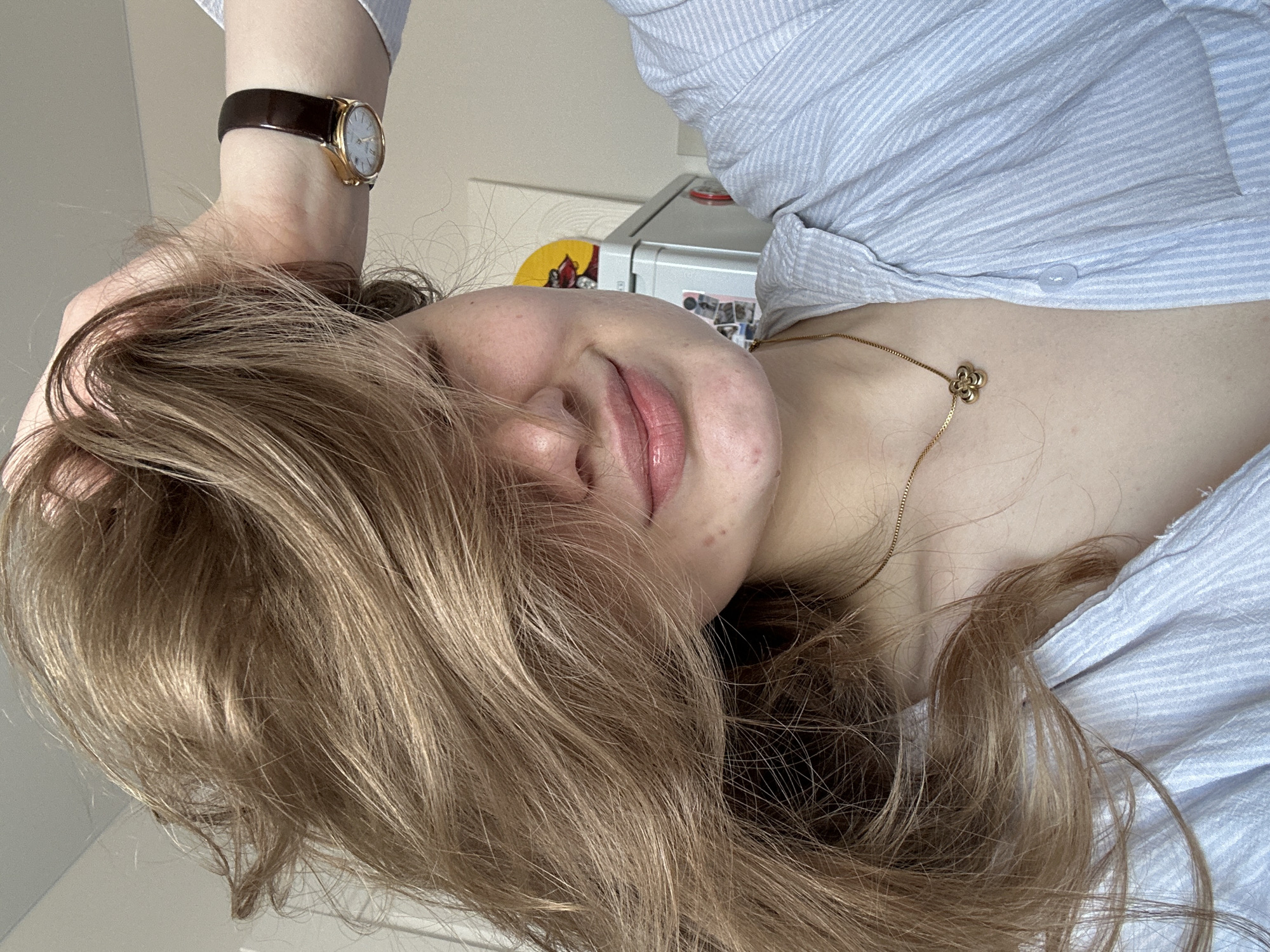 A woman with long, blonde hair is positioned in the frame. She is wearing a light blue, striped button-down shirt. A gold necklace with a flower-shaped pendant rests on her chest. A brown leather wristwatch with a gold face is visible on her wrist. The background contains a white wall and a metallic object, possibly a refrigerator. The location cannot be determined to be Kazan, Russia.