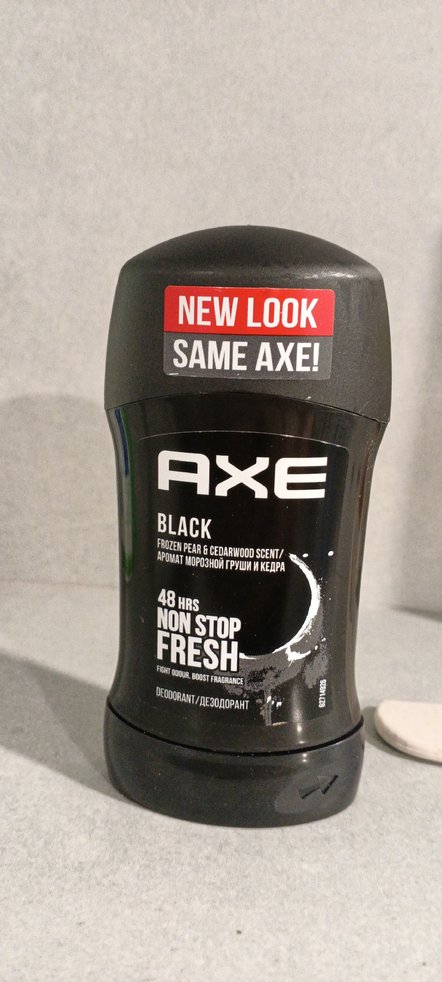 An AXE Black deodorant stick is centered in the frame against a light gray, textured background. The deodorant stick is black with white lettering. A red and white sticker on the top of the deodorant reads "NEW LOOK SAME AXE!". The main label displays "AXE BLACK" in large white font, followed by "FROZEN PEAR & CEDARWOOD SCENT/ АРОМАТ МОРОЗНОЙ ГРУШИ И КЕДРА". Below this, "48 HRS NON STOP FRESH" is prominently displayed, with smaller text "FIGHT ODOUR, BOOST FRAGRANCE" underneath. The bottom of the label reads "DEODORANT/ДЕЗОДОРАНТ". A small, partially visible white object, possibly a cotton pad, is present on the right side of the image. The image is a close-up, eye-level shot. There is no indication of the location within the city of Tashkent, Uzbekistan.