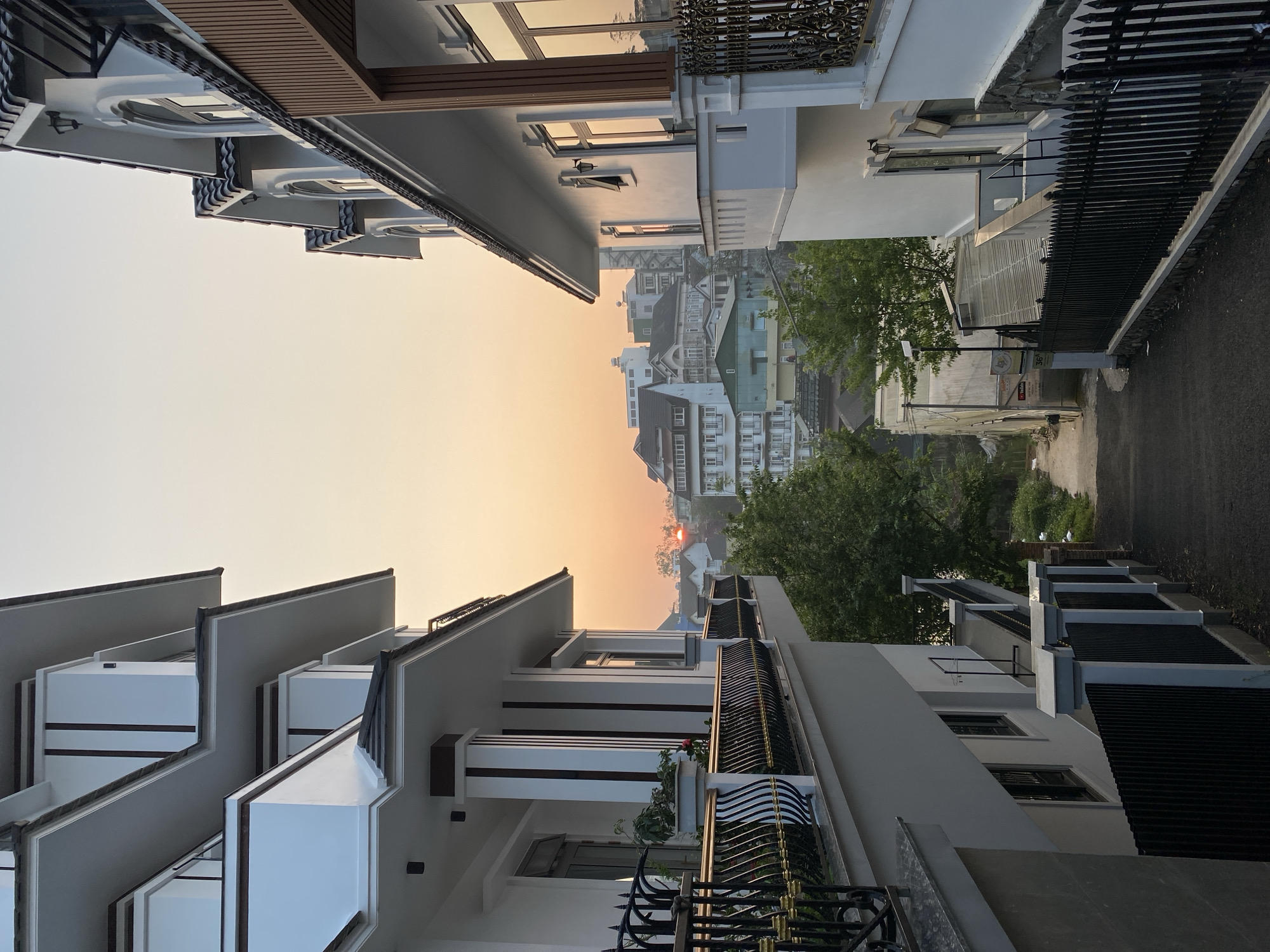 Captured in the urban area of Lâm Viên - Đà Lạt, Vietnam, the image showcases a street view with contemporary architectural structures lining both sides. These modern houses, predominantly white and gray, contrast with the warm tones of the setting sun visible in the background. The street extends into the distance, revealing more buildings and lush greenery. Dark wrought iron fences border some of the properties, adding a layer of depth to the composition. The atmosphere is serene, enhanced by the tranquil cityscape bathed in the soft, diffused light of either dusk or dawn.