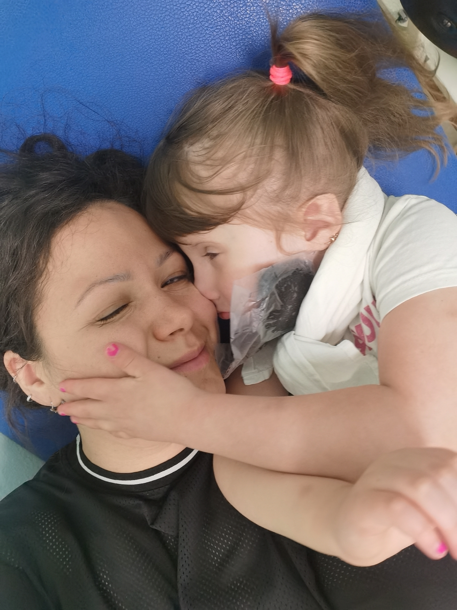 A medium-shot photograph captures a woman and a young girl lying down. The woman has dark, curly hair and is wearing a black mesh athletic shirt with a white-trimmed collar. The girl has light brown hair styled in a ponytail with a pink hair tie. She is wearing a white t-shirt and is leaning in to embrace the woman. The girl's arms are around the woman. Their heads are close together, and the woman appears to be smiling with her eyes closed. The background appears to be a textured blue surface. There is no discernible information about the image to suggest it was taken in Perm, Russia.