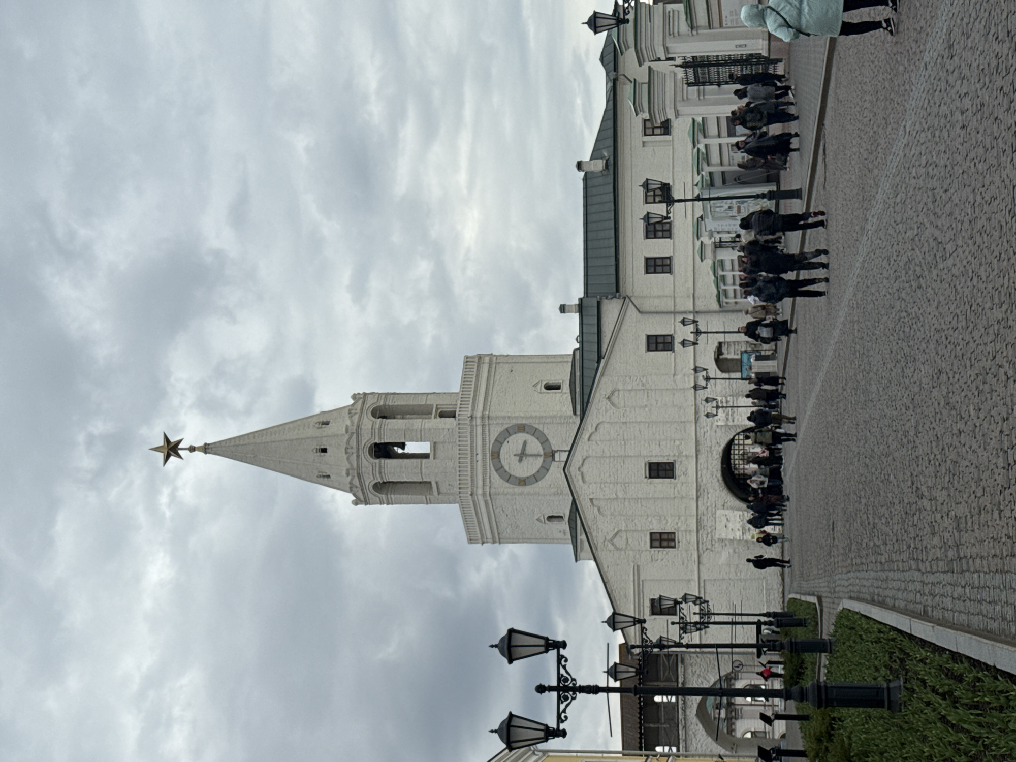 The image shows the Leaning Tower Syuyumbike in Kazan, Russia. The tower, made of white brick, features a clock face and a gold star on its spire. The sky is overcast. People walk on a stone-paved pathway. Ornate street lamps line the pathway. Adjacent is a strip of green vegetation.