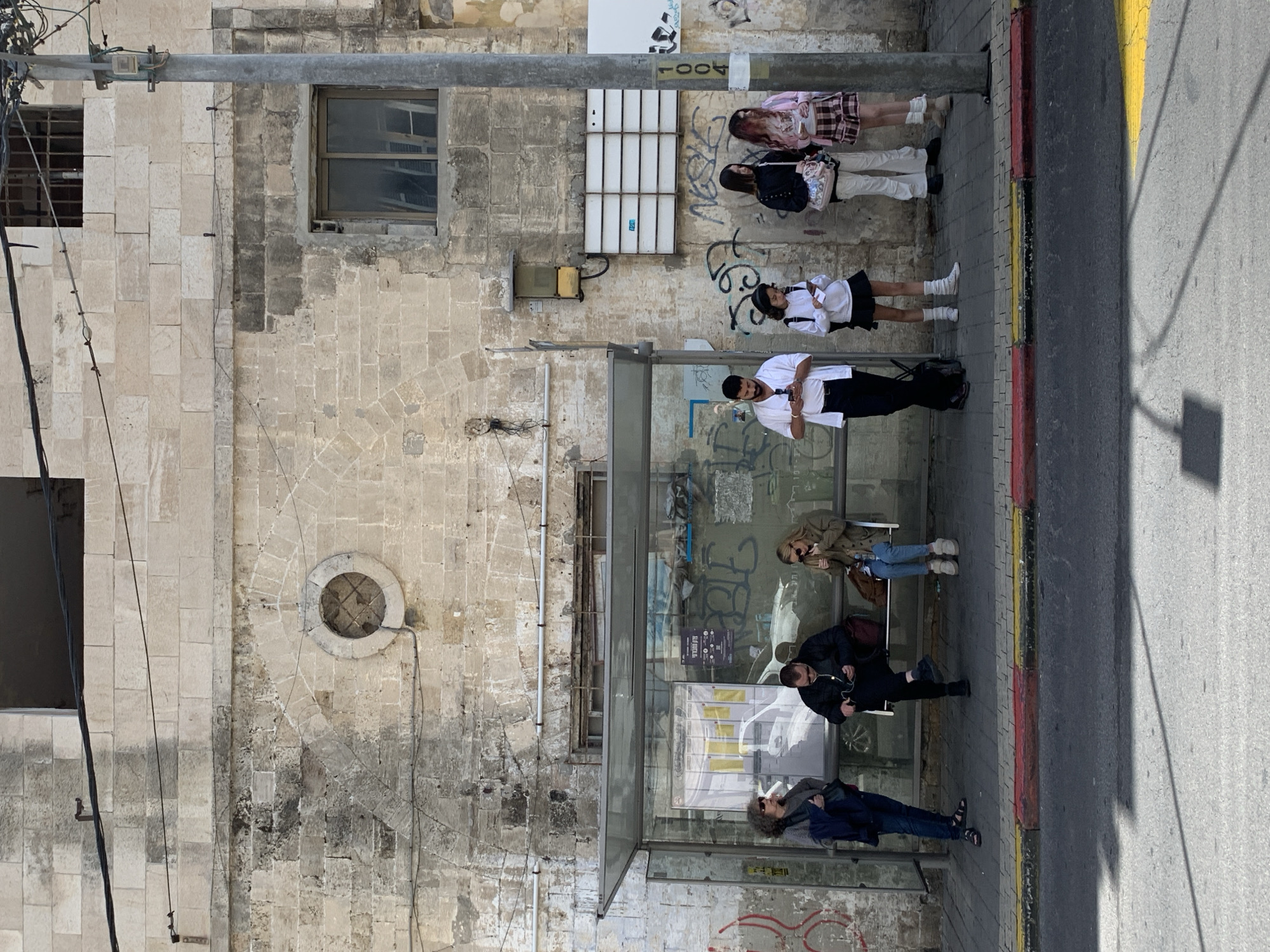 The image captures a street scene in Tel-Aviv, Israel. Along a sidewalk, a bus stop provides shelter for several people. Three individuals are seated on the bus stop bench. An additional person is standing outside the bus stop, focused on a mobile phone. Nearby, a group of three people are standing and engaged in conversation. The background features an aging building with architectural details, including multiple windows and a circular opening. The building's stone exterior shows signs of wear. Graffiti is visible on the bus stop structure and the lower portion of the building. A street and its shadow are visible beside the sidewalk. The overall scene appears to be captured during daylight hours.