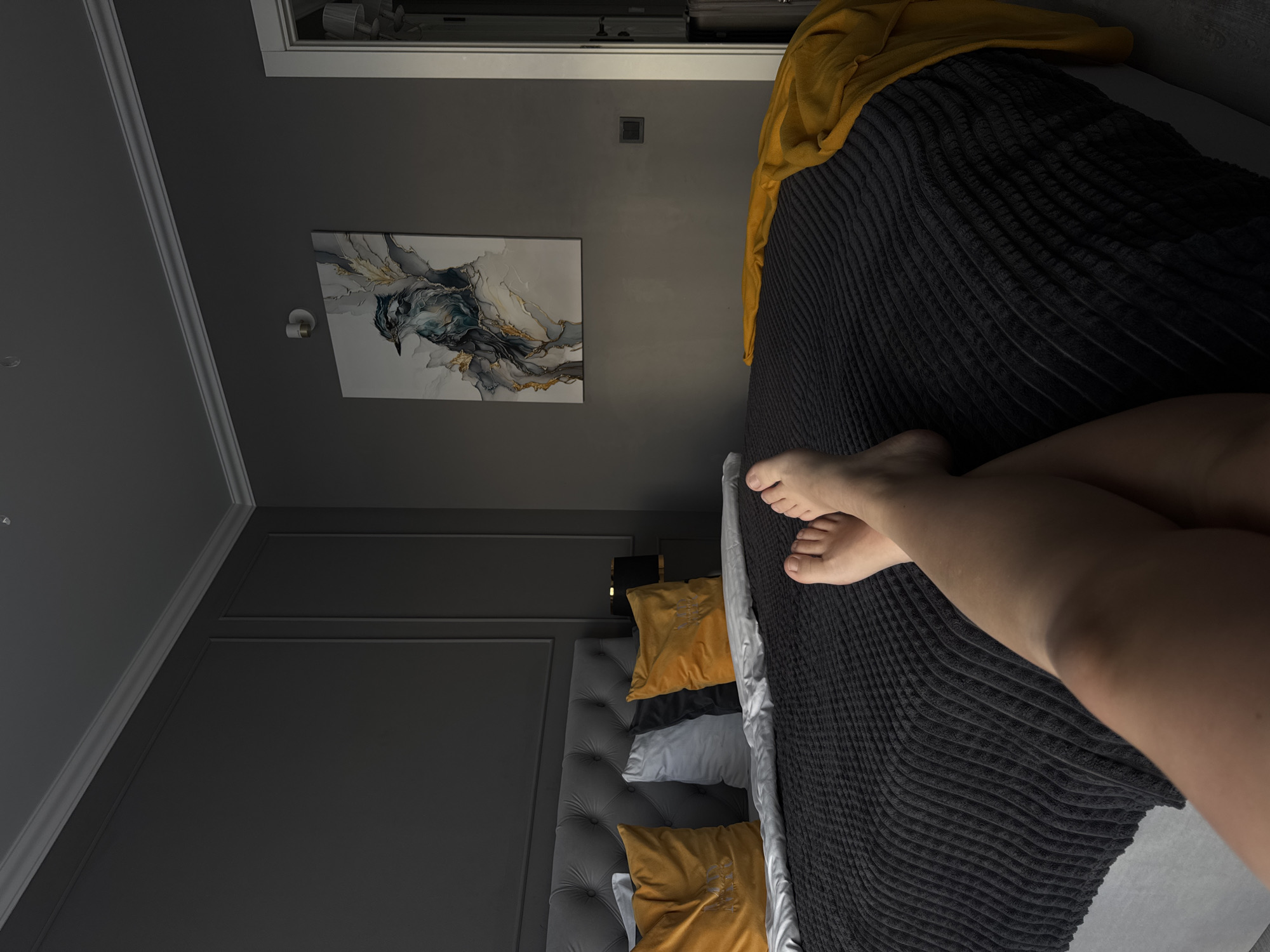 A bedroom interior is presented in a high-angle shot. A black, corduroy-textured bedspread covers the bed, with the lower right corner of the frame occupied by a person's legs and feet. The headboard of the bed is upholstered in gray and adorned with button tufting, and a set of pillows in shades of yellow, white, and gray are visible against it. A piece of abstract art hangs on the gray wall above the headboard. The upper right corner of the bed is covered with a yellow piece of clothing. The location is most likely in Volgograd, Russia.