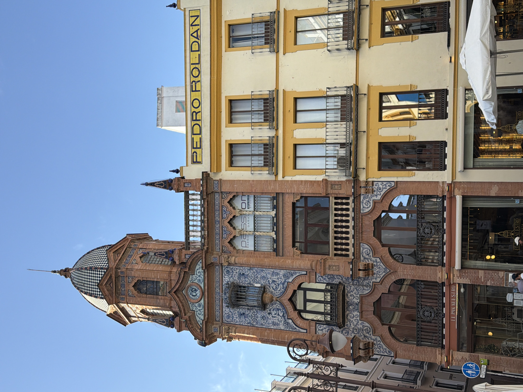 The image depicts a street scene in Sevilla, Spain, featuring two adjacent buildings with distinct architectural styles. The building on the left is ornate, with a domed tower topped by a spire and decorated with blue and white tiles and intricate brickwork. It has arched windows and balconies with decorative metal railings. Below the upper floors, a sign reads "EL PAN NUESTRO" above the entrance to a shop.
The building on the right is painted a light yellow and has a more modern, rectilinear facade. It features multiple floors with regularly spaced windows, each with a balcony and dark metal railings. A large yellow sign with black lettering across the top reads "PEDRO ROLDAN." The windows on this building reflect the surrounding environment, showing other buildings and the sky.
In the foreground, on the left side of the image, a decorative lamppost is visible. Towards the bottom right, a white umbrella shades an outdoor seating area, suggesting a cafe or restaurant. Several people are visible on the sidewalk in front of the shops, and a traffic sign is present on the far left. The sky is clear and blue. The overall impression is of a vibrant urban environment with a blend of historical and contemporary architecture.