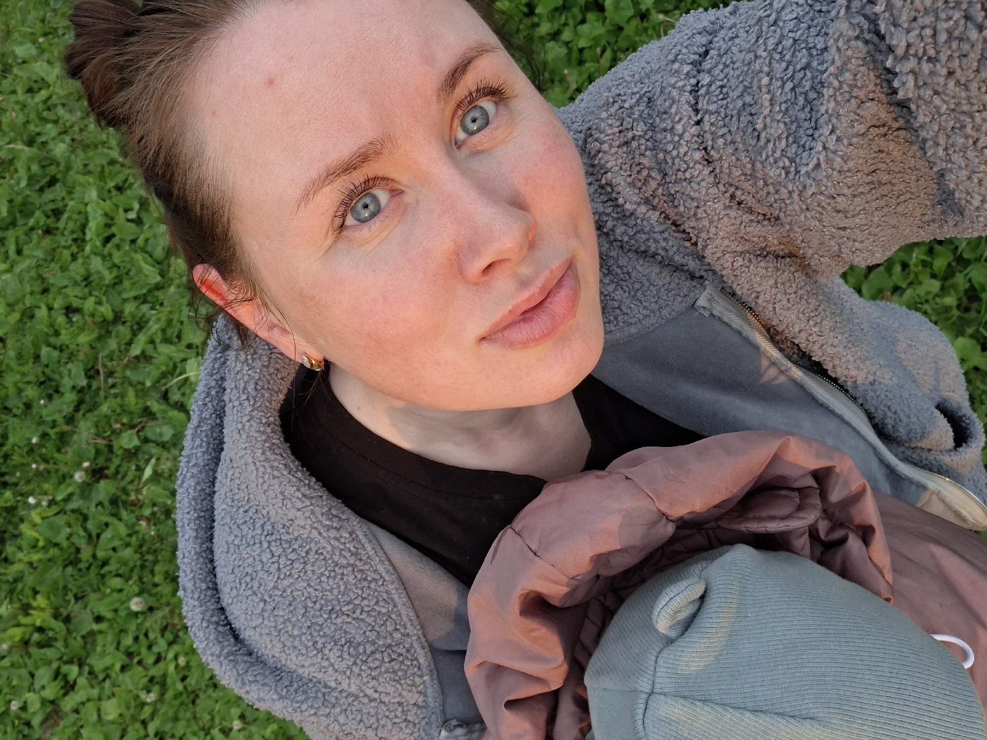 A high-angle, medium shot captures a young Caucasian woman from the shoulders up, lying on a patch of green grass. Her eyes are open and looking up at the camera, and her mouth is slightly parted. She has light skin with a rosy flush, fair eyebrows, and long eyelashes. Her light brown hair is pulled back into a bun. She is wearing a gray, fuzzy hooded jacket over a black shirt, and holding a bundle of clothing in a blush pink and light blue color. The background consists of green grass with scattered small white flowers, suggesting an outdoor setting possibly in a park or field within an unknown city. The lighting suggests it is late afternoon or early evening, as indicated by the warm glow on her face.
