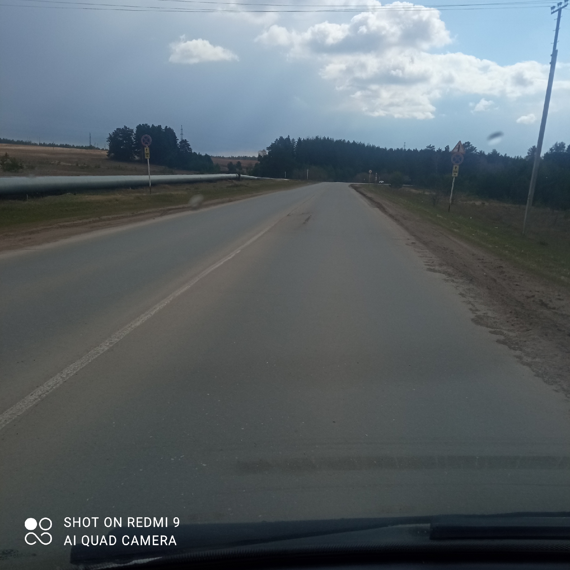 The image displays a straight asphalt road extending into the distance under a partly cloudy sky, taken from inside a vehicle with the dashboard visible at the bottom.
On the left side of the road, a large, light-colored pipeline runs parallel to the asphalt. Beyond the pipeline lies a field of dry, light-colored grass, backed by a dense forest of dark green coniferous trees. A cluster of road signs is visible: a yellow diamond-shaped sign with an 'X' inside (indicating a crossroad or intersection ahead), below it a circular red sign with a diagonal line (typically meaning "No Stopping" or "No Standing"), and further below, a yellow rectangular sign with "100 M", likely denoting a distance. In the far distance, a tall power line tower can be seen against the sky.
On the right side of the road, a similar dense forest lines the route. Road signs are also present here: a yellow diamond-shaped sign with an 'X' (crossroad ahead) and beneath it, a yellow rectangular sign with "100 M". A tall wooden utility pole stands on the right. In the far distance on the right, a small white vehicle is faintly visible on the road.
The asphalt road shows some signs of wear, with minor cracks or patches in the surface. A solid white line marks the left edge of the visible lane. Overhead, power lines span across the sky. The sky itself is a mix of bright blue with scattered white cumulus clouds, along with darker, more subdued cloud formations, suggesting a mix of sunny and overcast conditions.
In the bottom-left corner, a white watermark states "SHOT ON REDMI 9" and "AI QUAD CAMERA". The scene depicts a rural or semi-rural landscape, indicative of a journey through the Прикамский region of Russia during daytime.